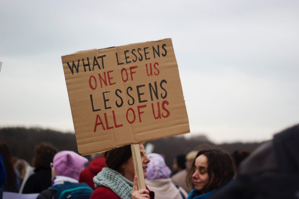 A protestor holds a sign saying "what lessens one of us, lessens all of us"