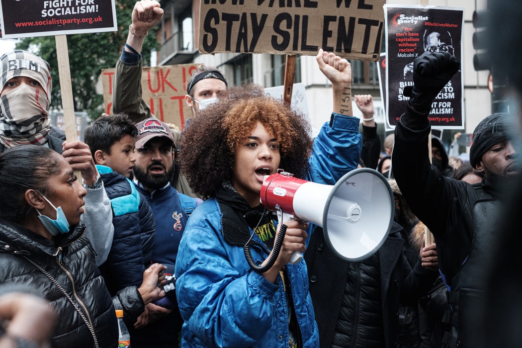 A black woman speaks into a megaphone with her fist in the air. She is surrounded by other protesters