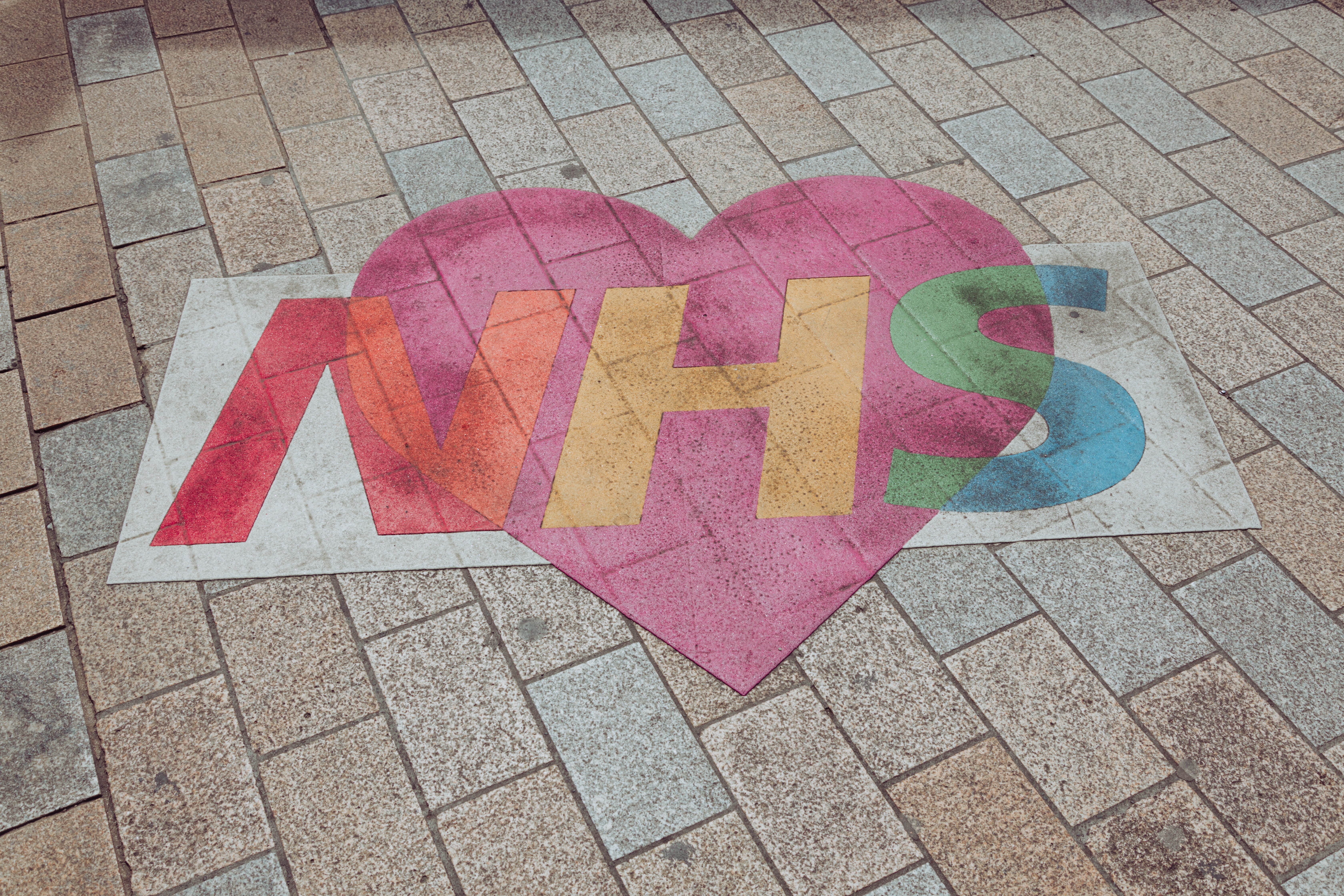 A chalk drawing of the NHS logo in pink, yellow and green lies on top of a red heart on the pavement. 