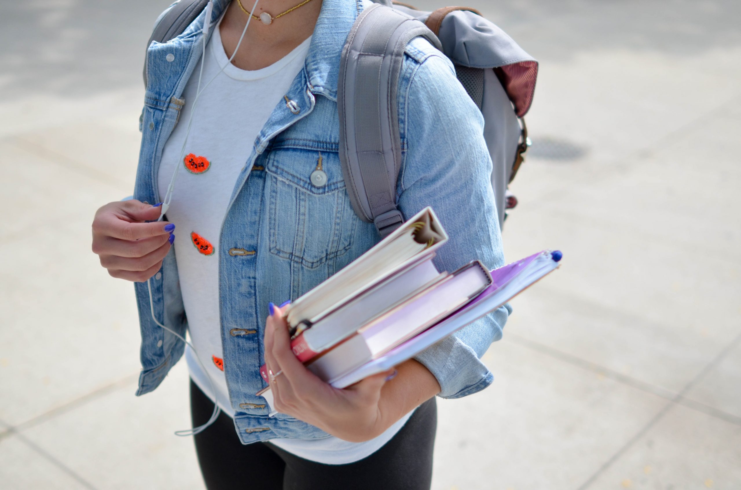 A white university student stands diagonally on to the camera, while wearing a denim jacket. Her right hand holds the edge of her jacket, her left arm holds her notebooks and books for classes. 
