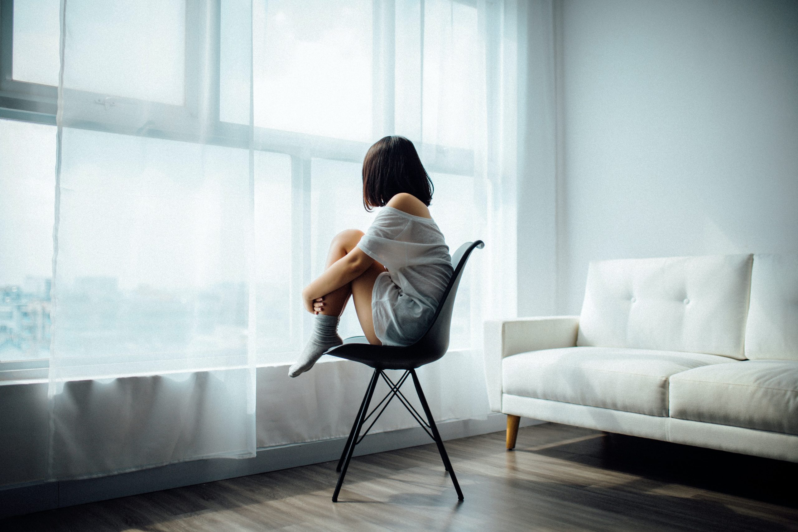 A young girl sits on a black chair in a nearly all white room. She is clutching her knees to her chest and staring out the window. She is wearing white and has black hair cut into a bob. 