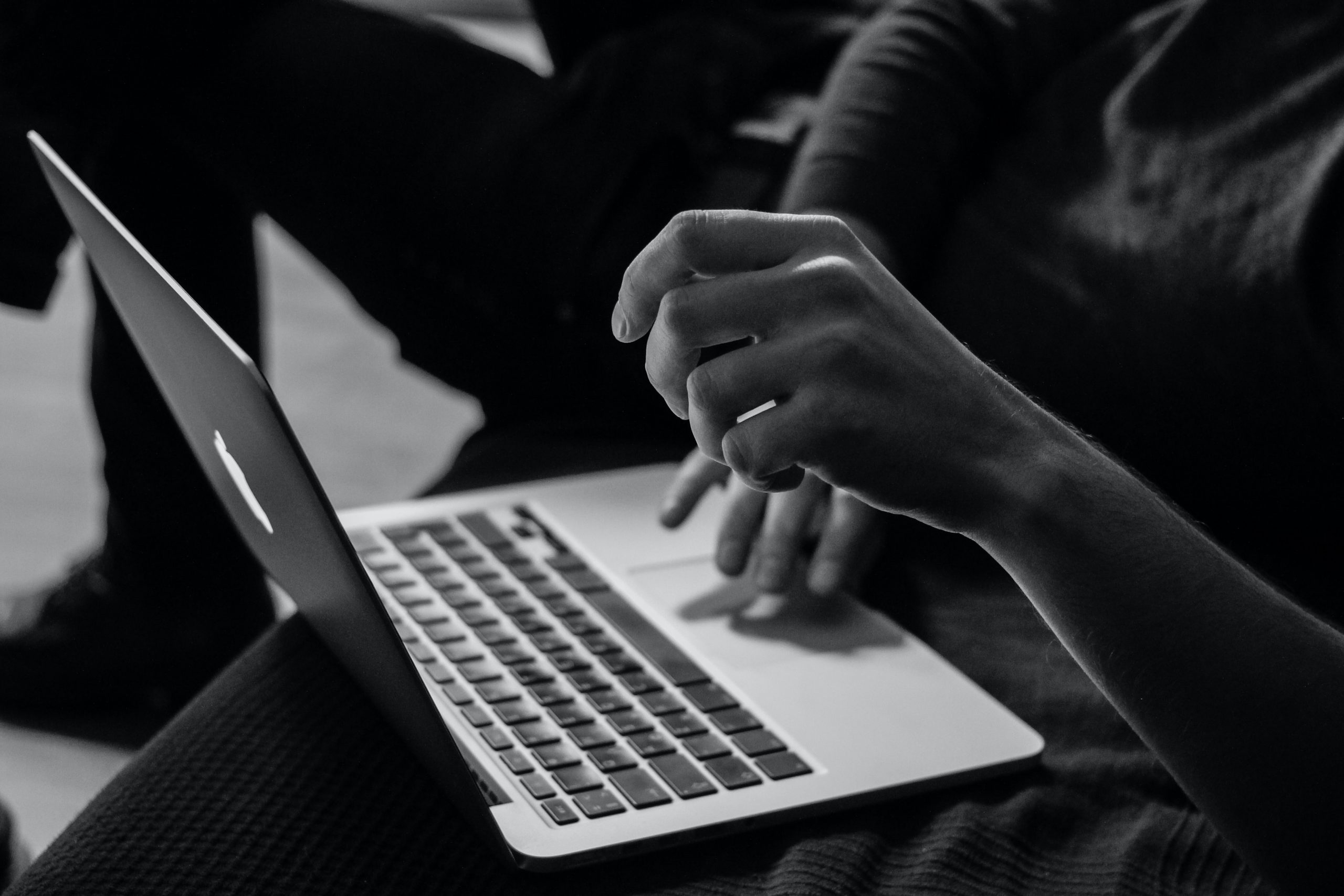 An monochrome image shows two black hands typing on a MacBook laptop. 