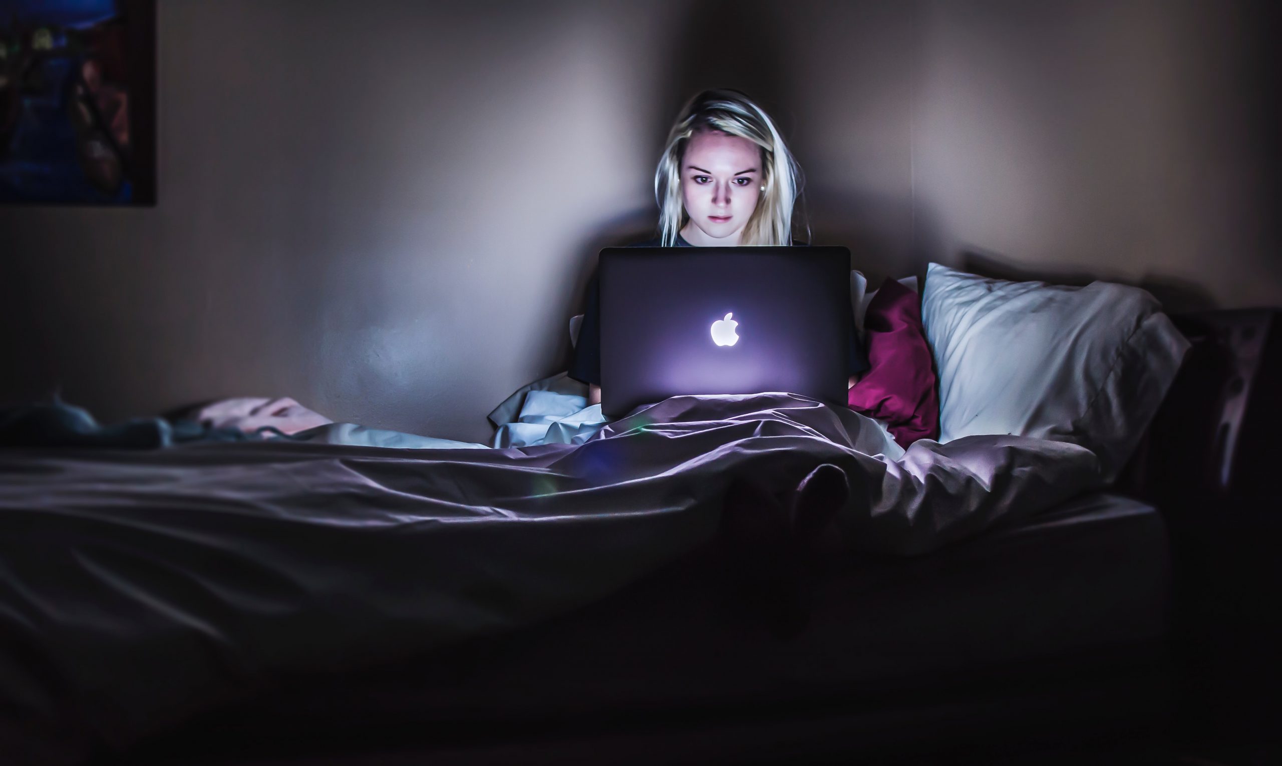 A white woman with blonde hair sits on her bed in the dark. Her face is illuminated by her macbook screen.