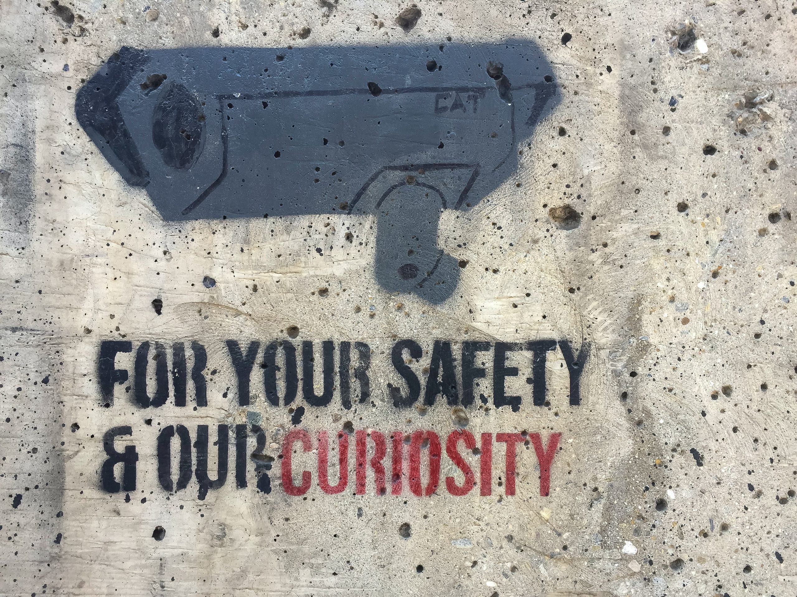 Graffiti on a beige wall shows a grey security camera with the words "for your safety and our curiosity" underneath