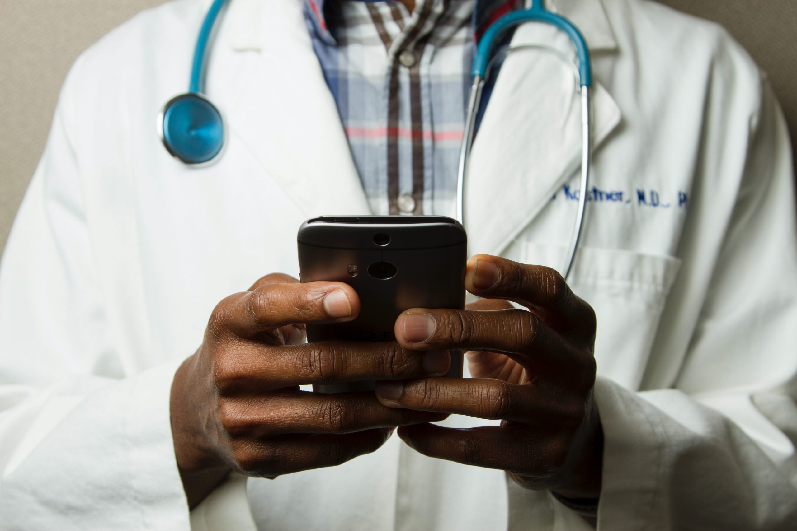 The photograph shows the chest and hands of a black doctor holding a mobile phone