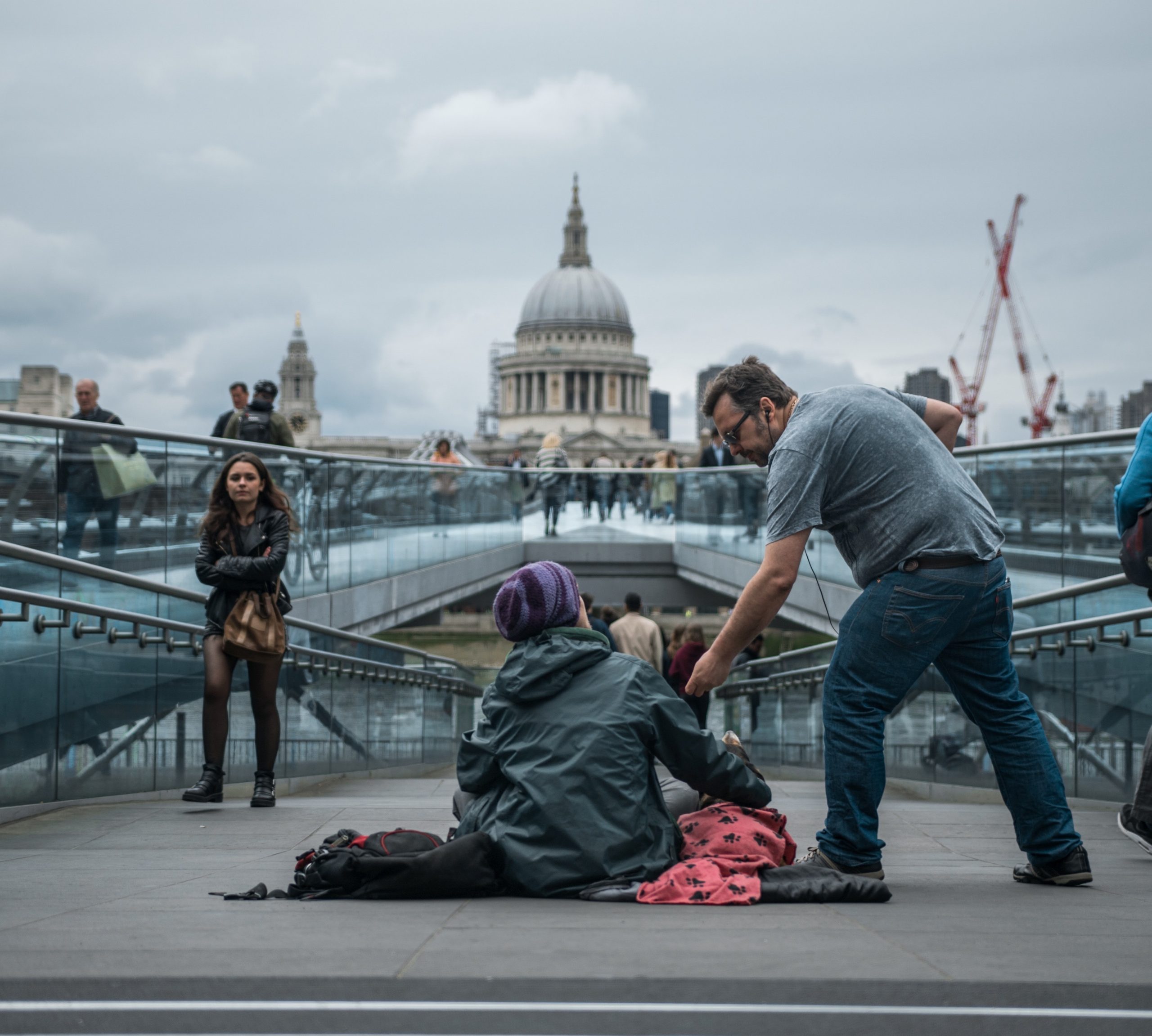 A homeless man sits on the end of London Bridge. He looks up while a passerby hands him some change.