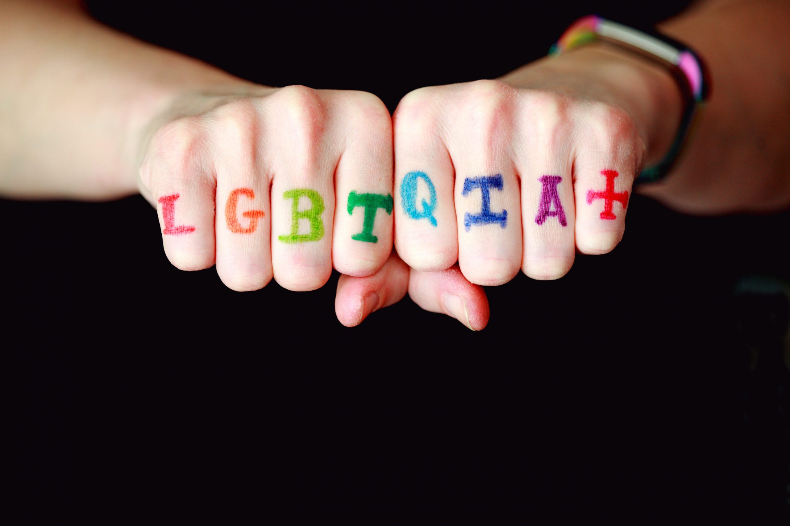 A pair of closed fists shows "LGBTQIA+" written in rainbow colours across the knuckles 