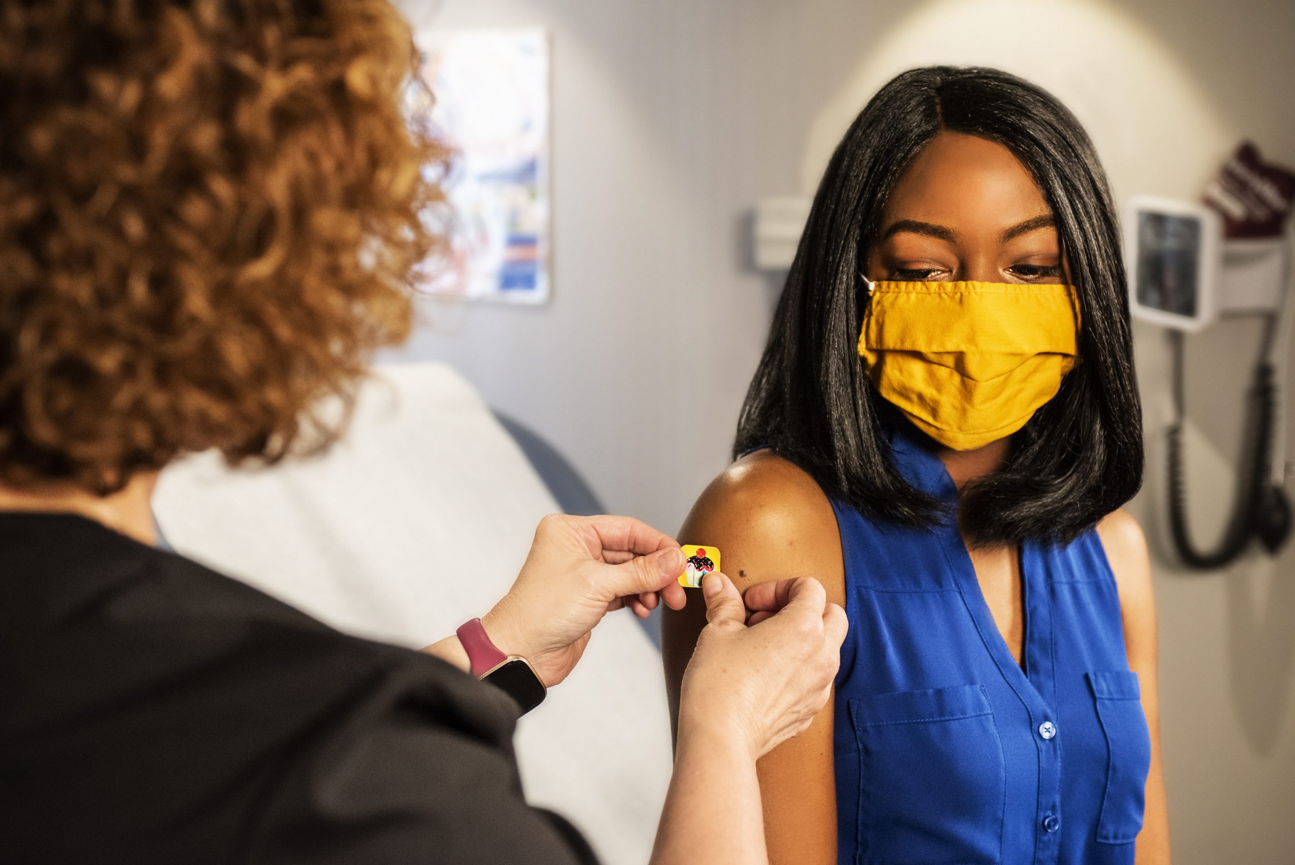 A black woman wearing a yellow face mask and blue top is given her covid vaccination by a person wearing a black top with curly hair. 
