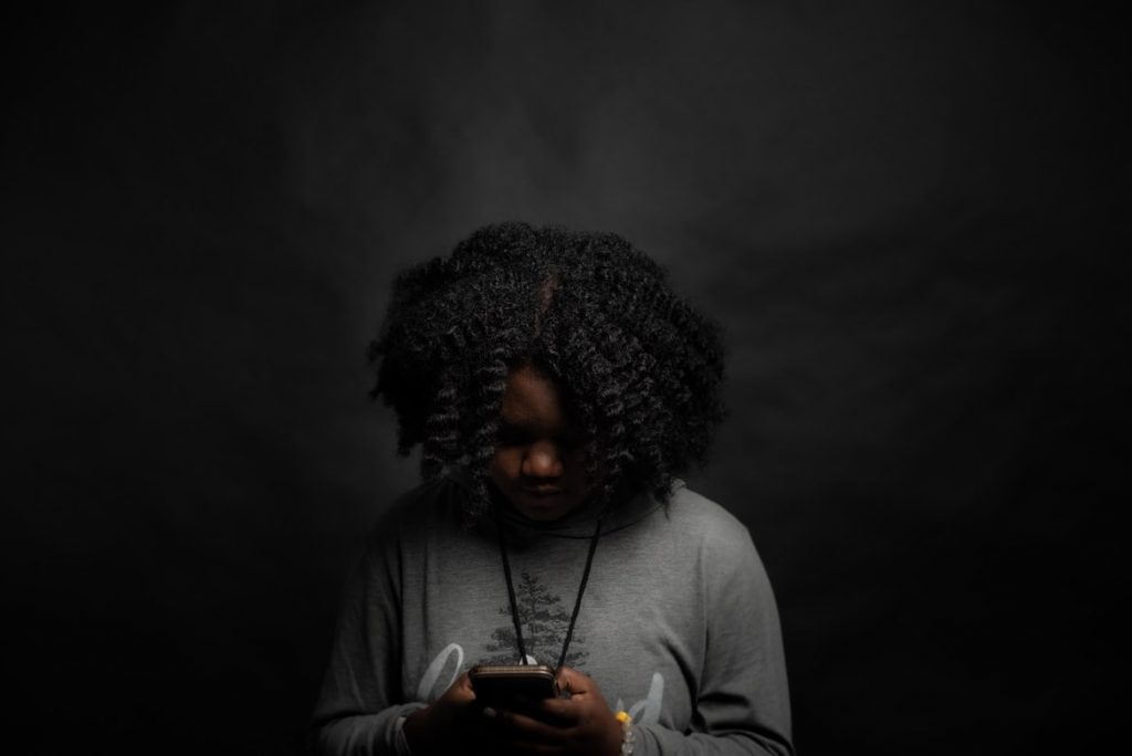 A Black woman in a grey top looks down at her mobile phone in a dark room