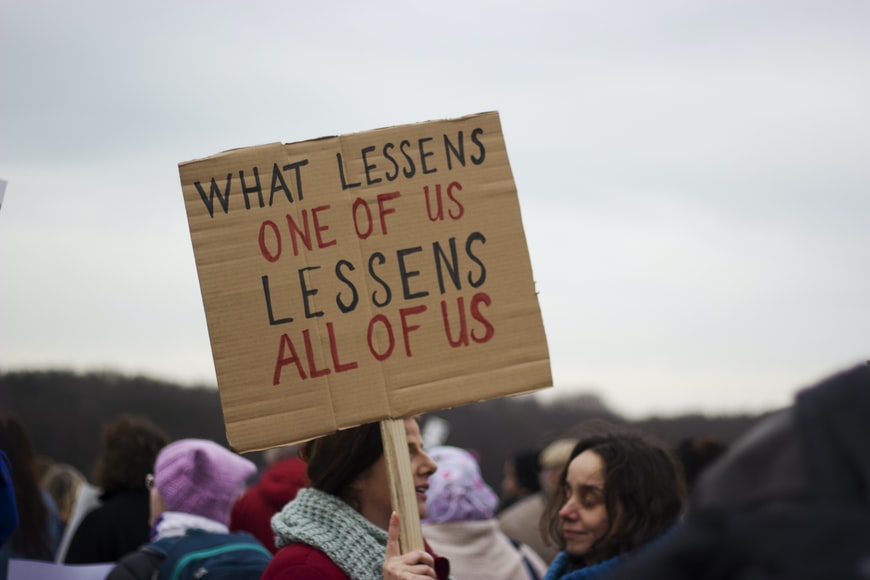 A placard held aloft by protesters reads 'What lessens one of us lessens all of us'