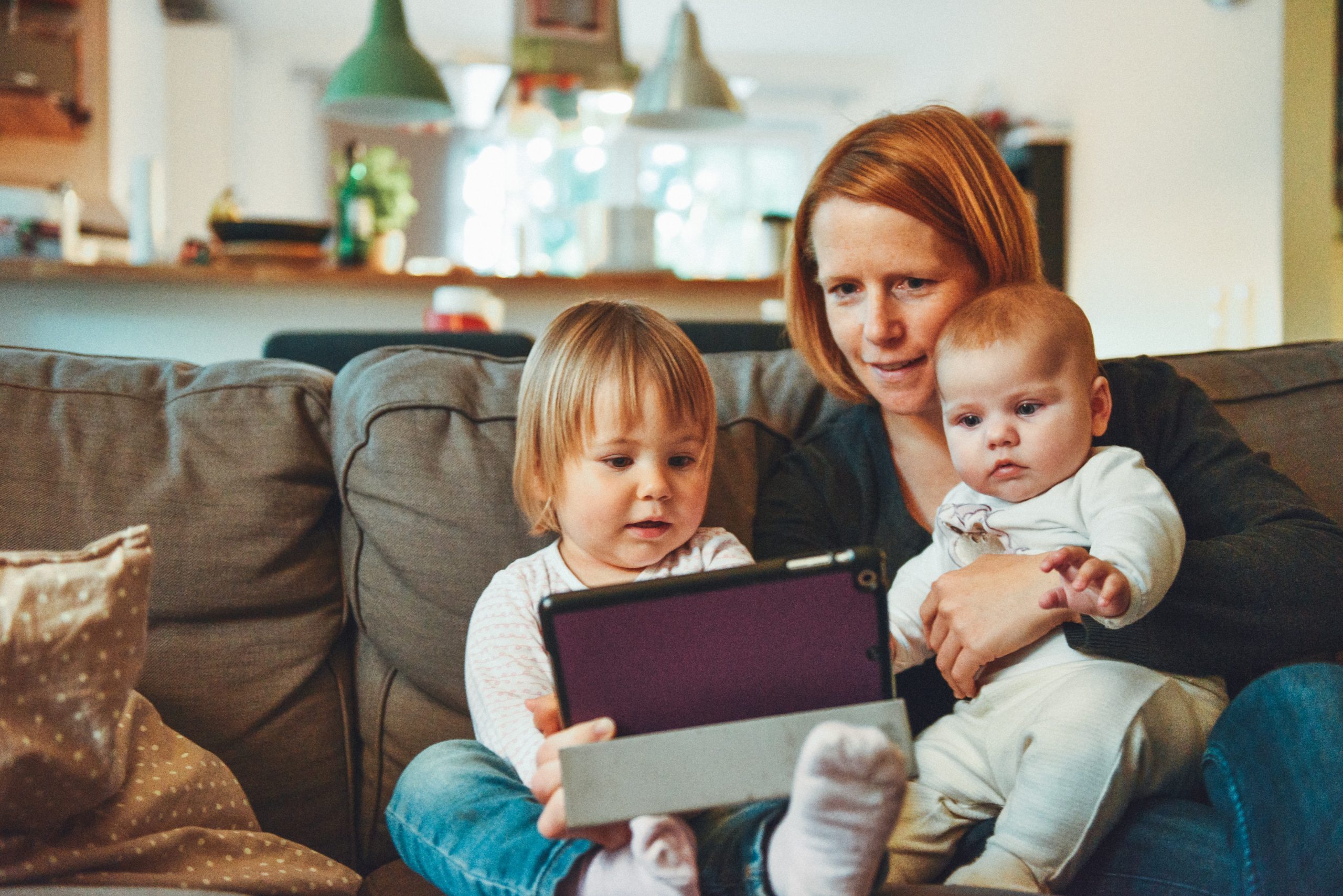 A young white mum cradles a toddler and a baby in her lap. She sits on a sofa with a tablet in her hands in front of the children.