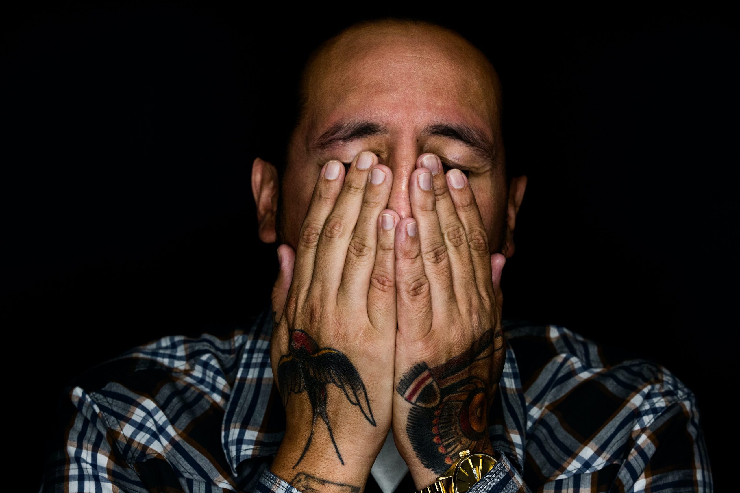 A man with swallow tattoos on his hands clasps them to his face in despair. He is wearing a blue checkered shirt against a black background.
