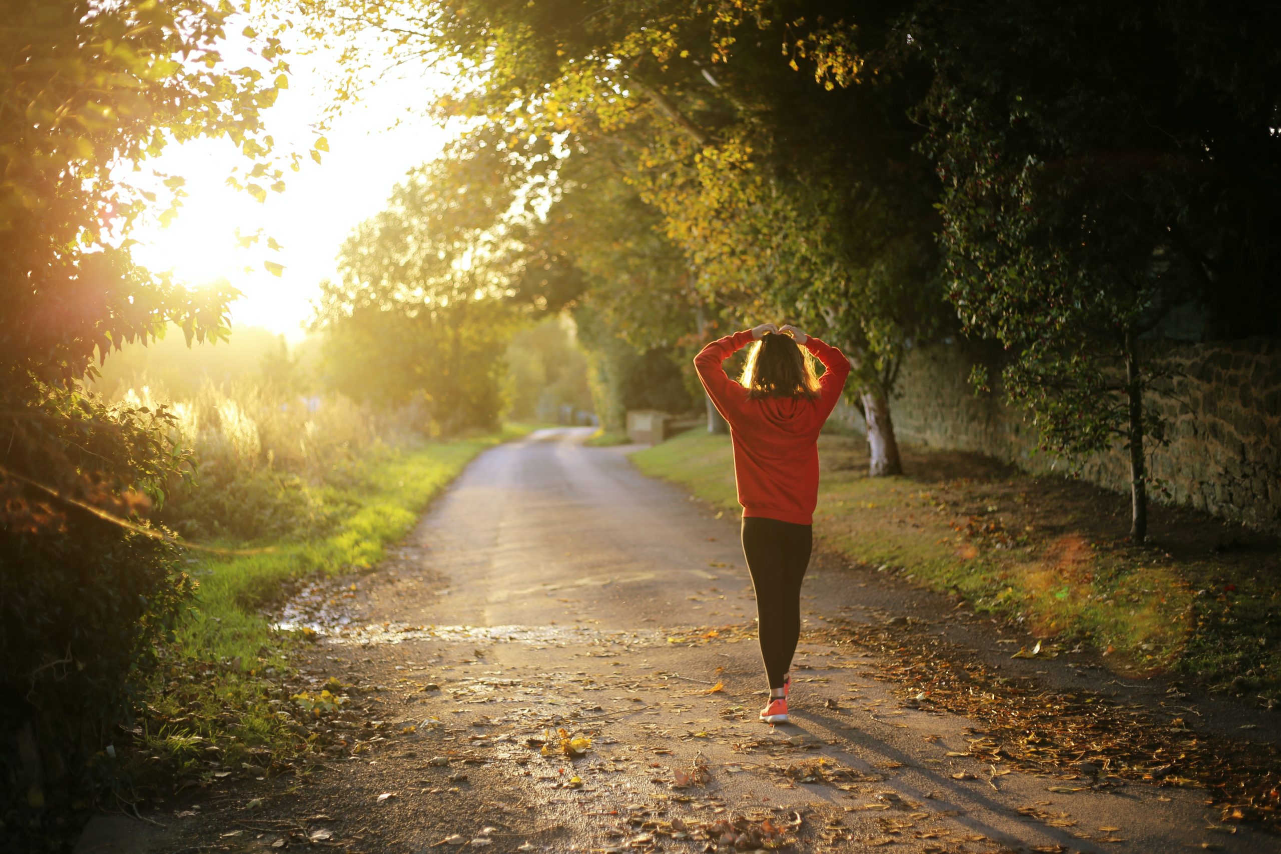 A woman wearing a red long sleeved top and black trouseres walks away from the camera with her hands on her head. She is walking down a path at golden hour between some trees.