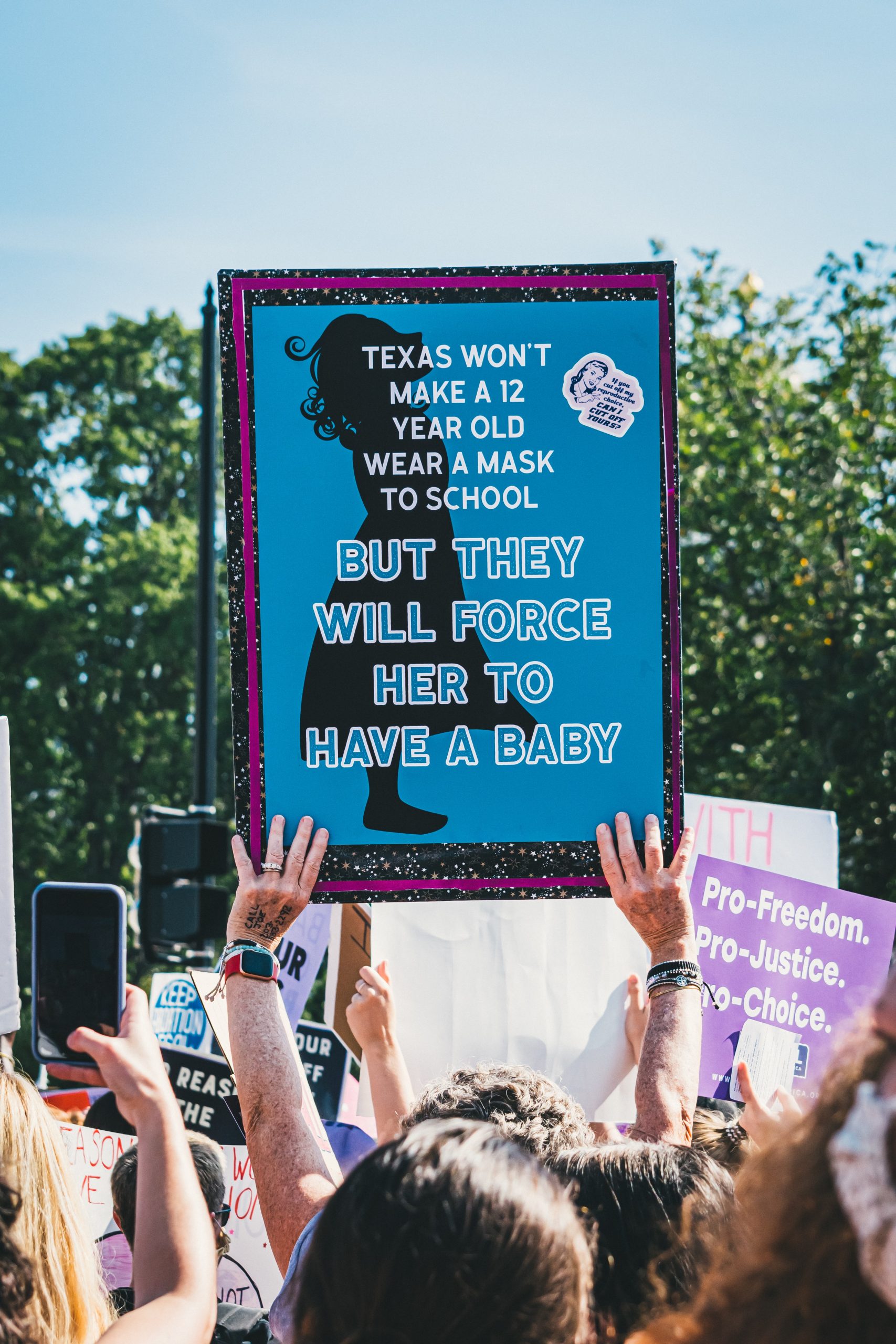 A sign his held above a crowd of protesters. I shows a young girl silhouetted in black on a blue background. The text reads: "Texas won't make a 12 year old child wear a mask to school but they will force her to have a baby"