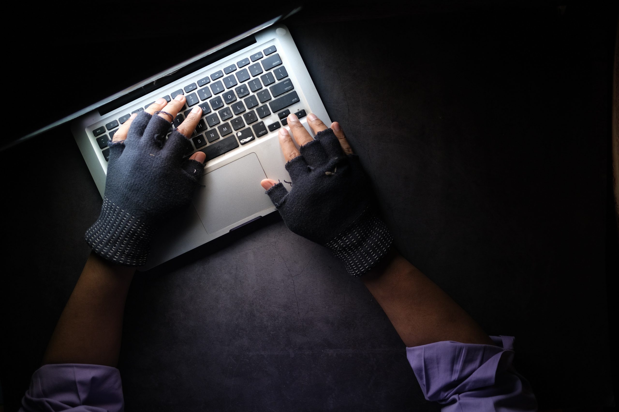 An aerial shot shows a pair of hands wearing gloves typing on a Macbook laptop. The photo is lit only by the laptop screen. 