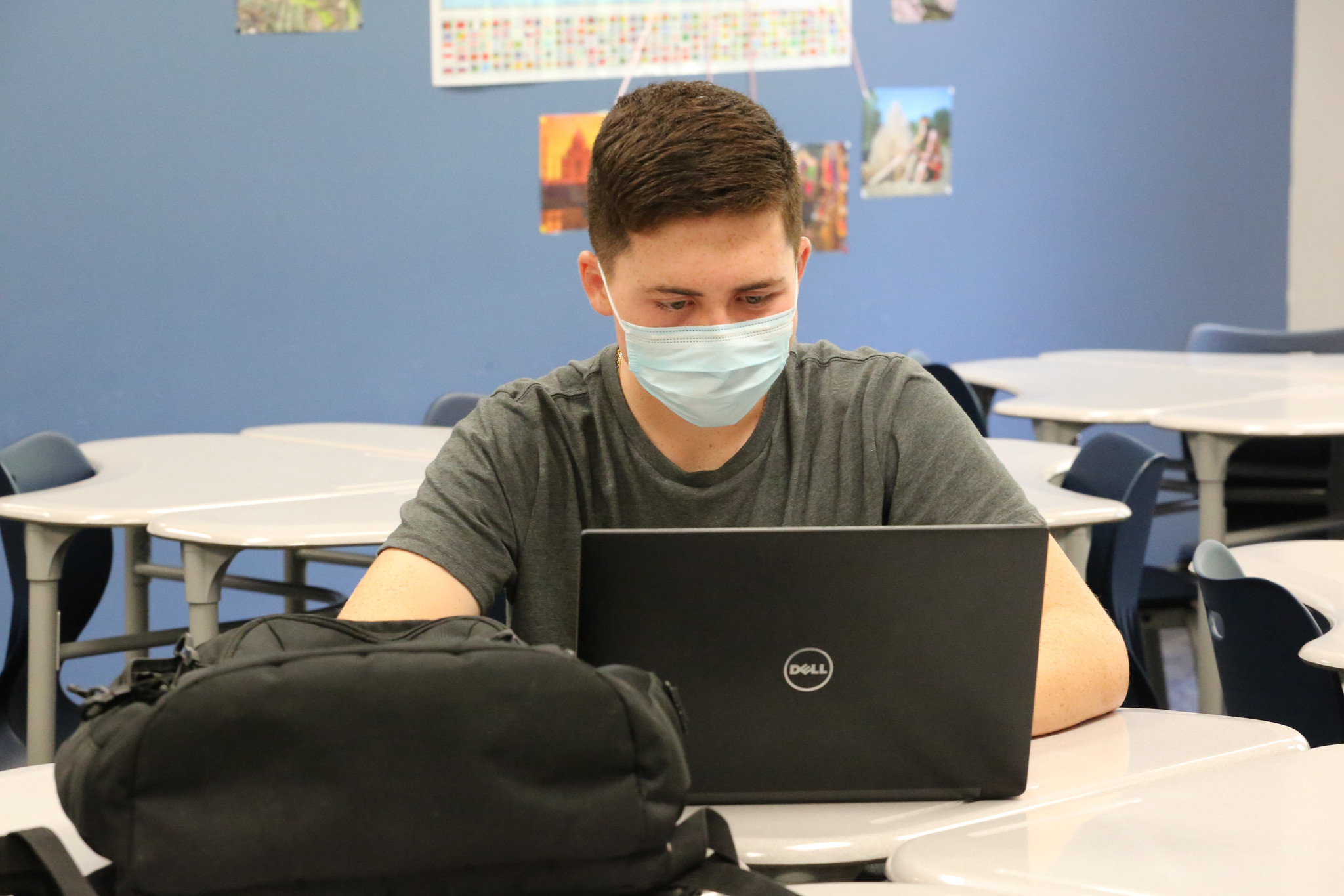 A young teenage boy sits at a desk on a laptop. He is wearing a grey t-shirt and a disposable face mask and has short, dark hair.