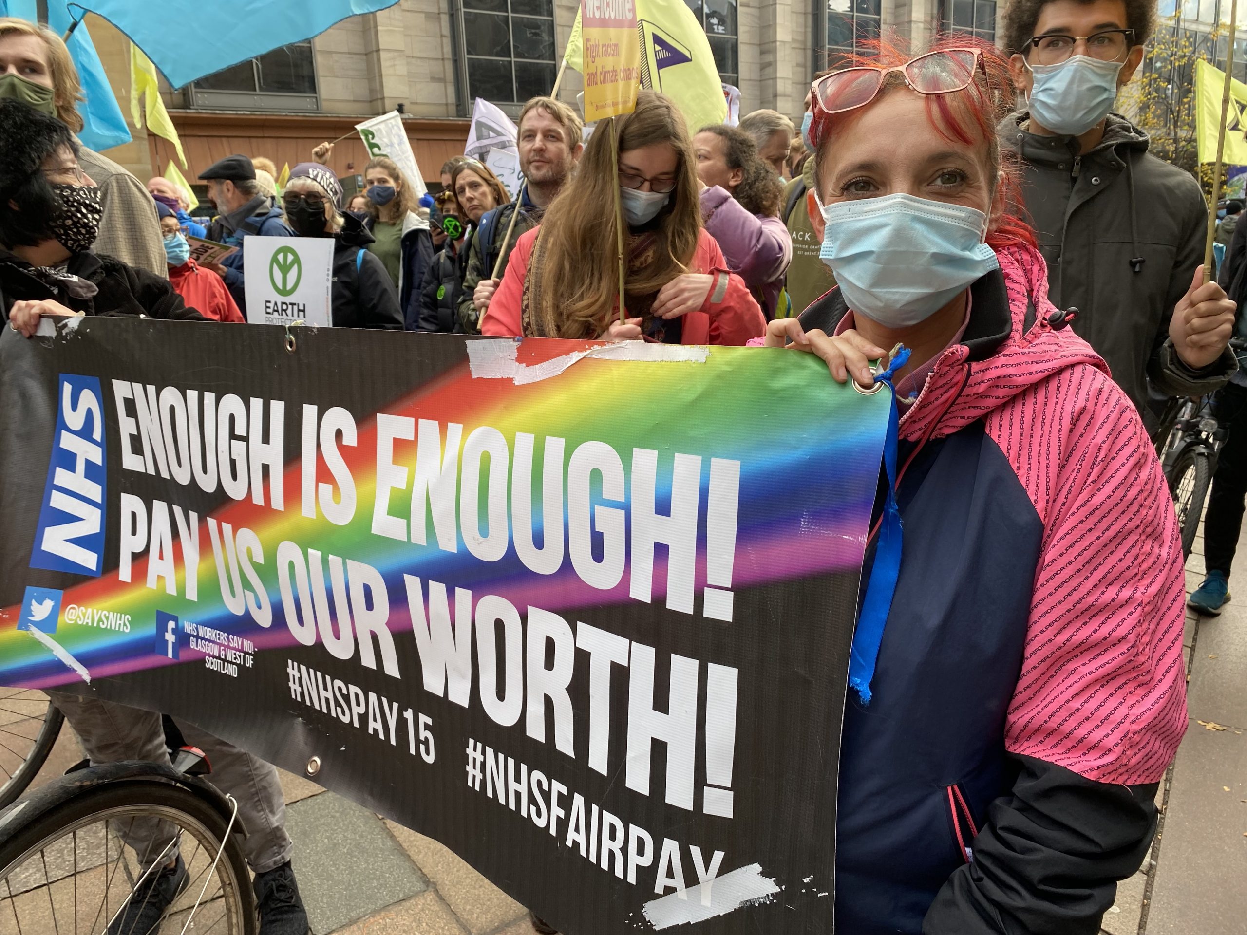 Brenda marches through the streets of Glasgow at the Extinction Rebellion demo, wearing a blue and pink jacket, glasses and facemask. She is carrying a banner which reads: 'NHS. Enough is enough! Pay us our worth!"