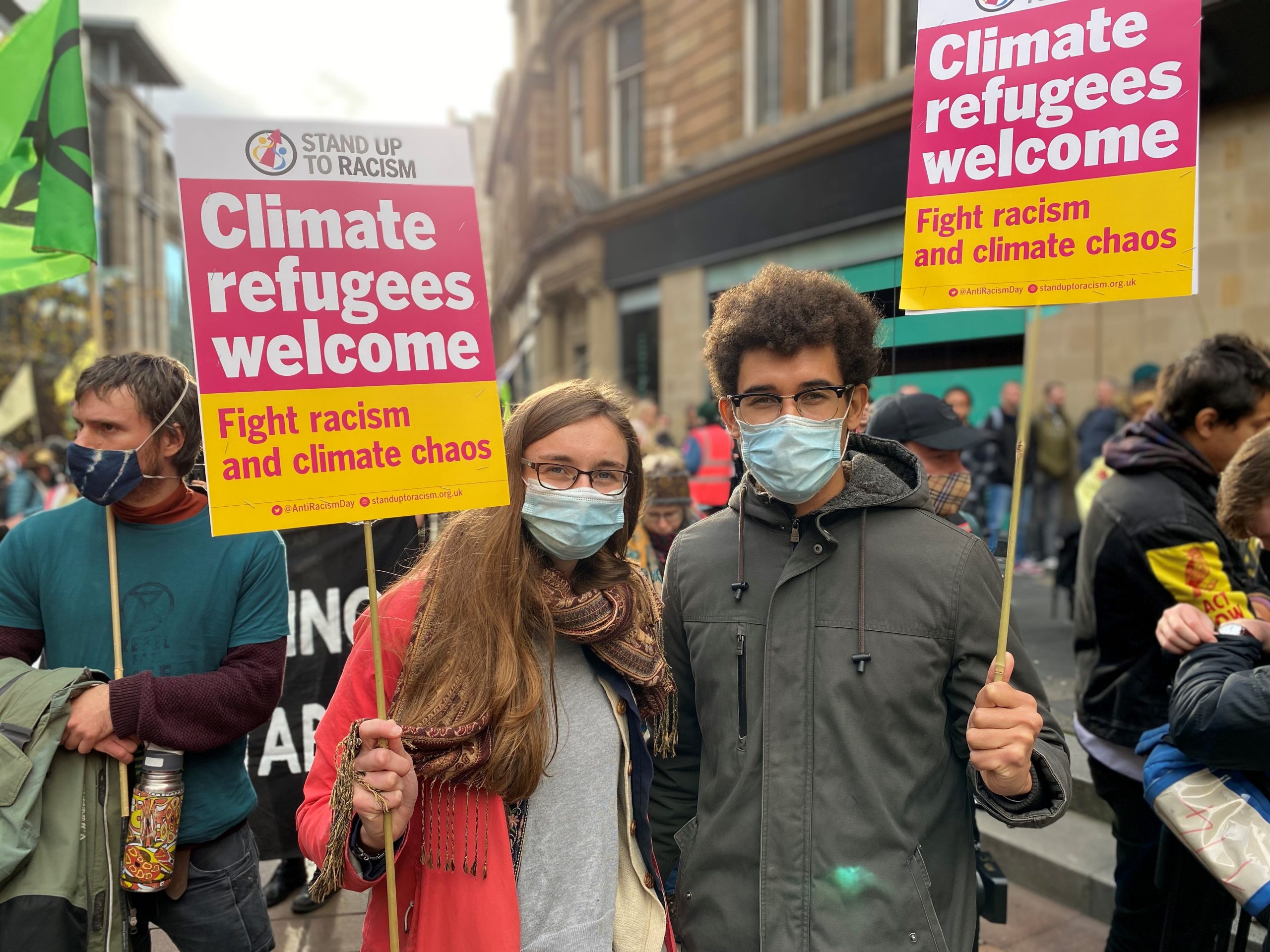 Protestors Joanna and Vladimir march together through the streets of Glasgow at the Extinction Rebellion anti-greenwashing demo, holding placards aloft which read 'Climate Refugees Welcome' - fight racism and climate chaos