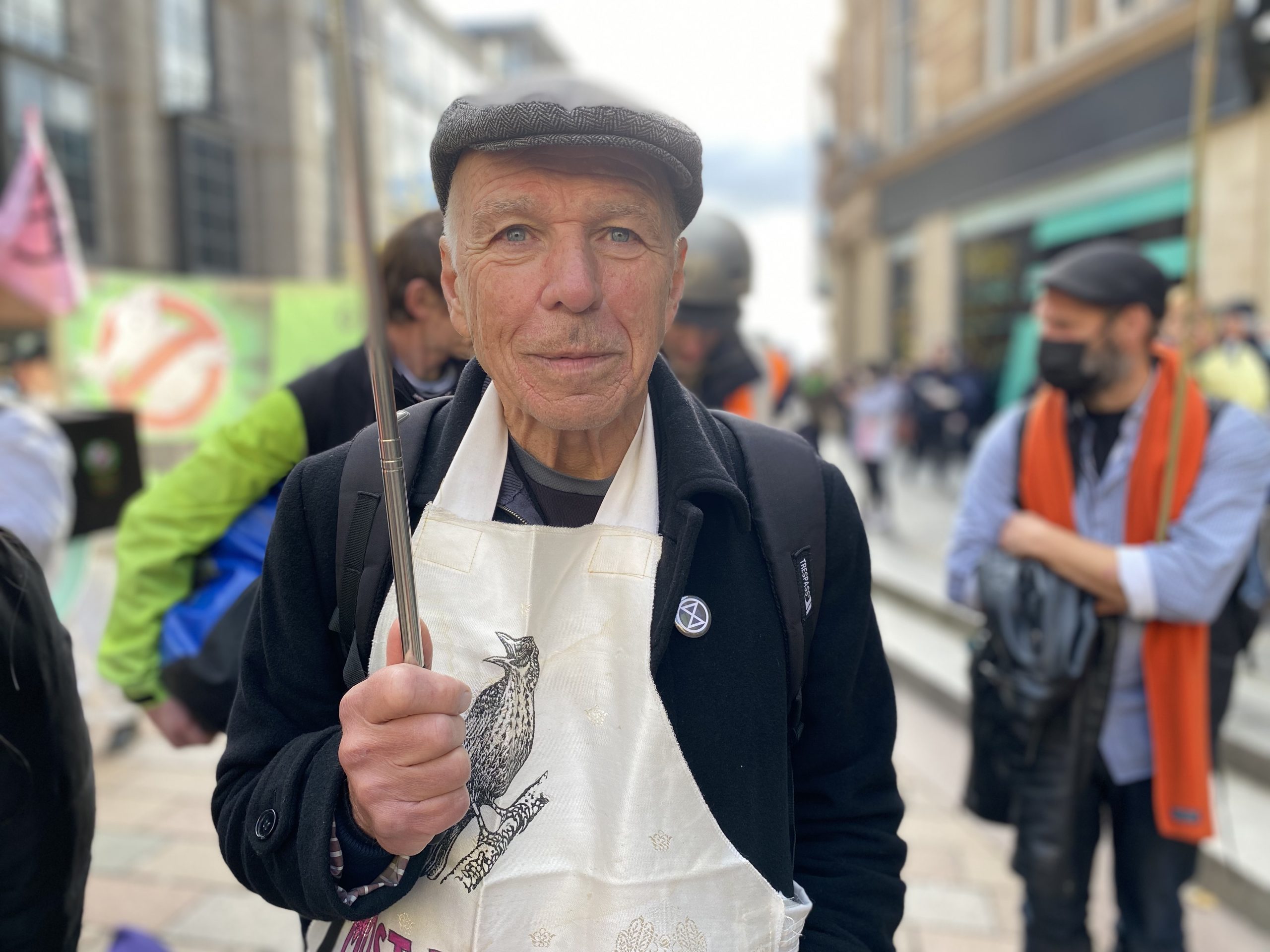 John walks the streets of Glasgow, taking part in the Extinction Rebellion demo. He is wearing a flat cap, a jacket with an XR badge on it and an apron with a picture of a bird on it.