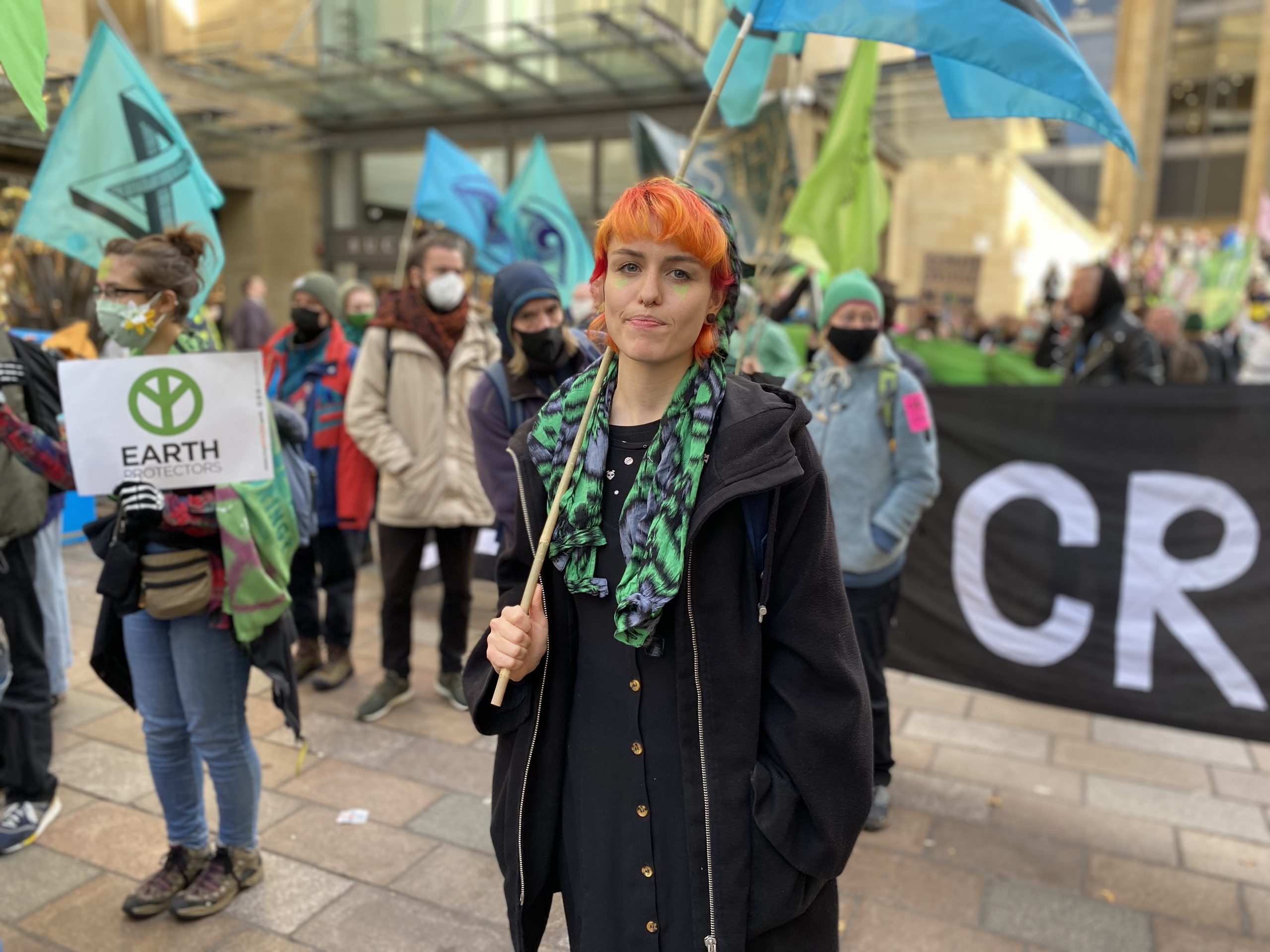 Reina stands amid fellow protestors at the Extinction Rebellion demo in Glasgow. She has orange hair, is wearing a jacket and scarf and is carrying a blue flag.