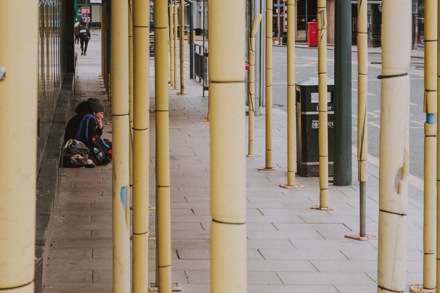 A homeless woman sits on a pavement near a litter bin amid scaffolding poles on a street in Leeds.