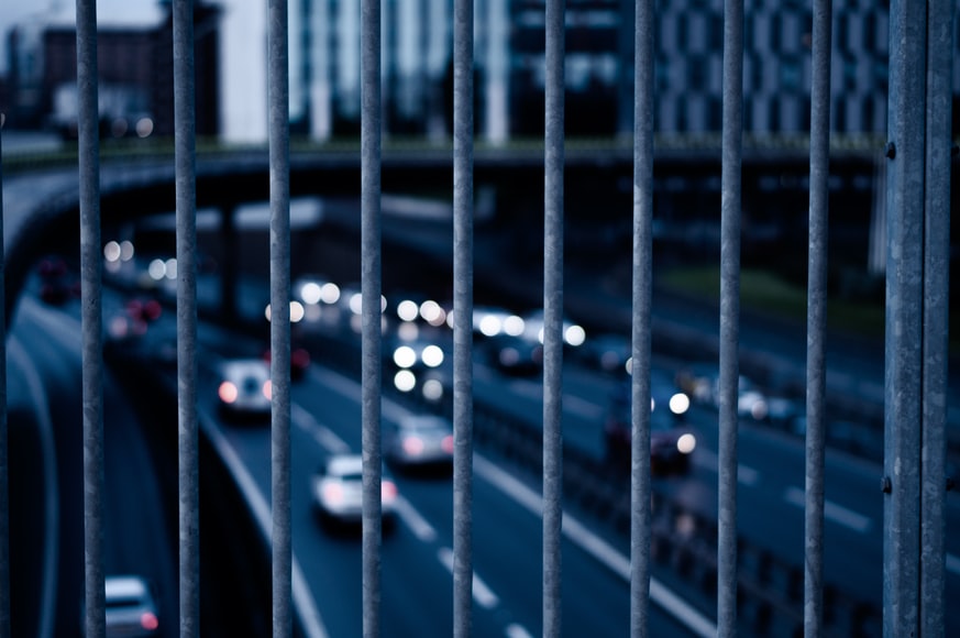 A motorway full of cars is photographed through the metal bars of a fence on a bridge above the road