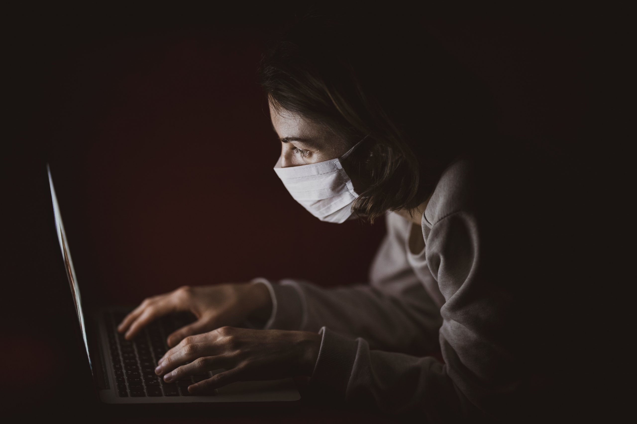 A person wearing a white face mask hunches over a laptop in the dark. The only light is coming from the laptop screen.