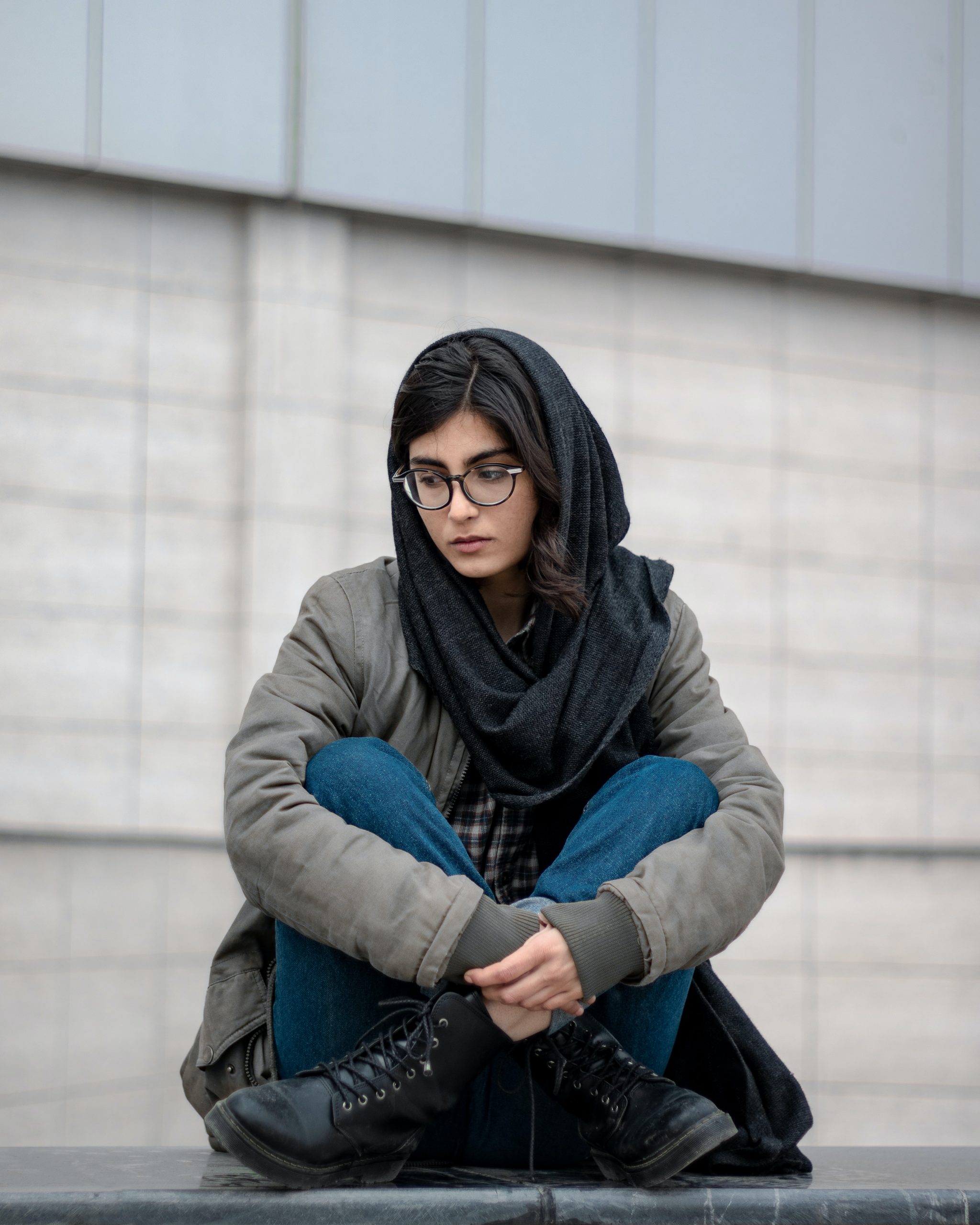 A woman wearing a hijabi sits cross legged on the floor looking pensieve. 