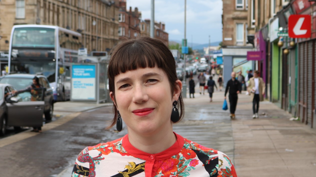Rhiannon J Davies stands on a pavement looking at the camera in a multi-coloured top with a double-decker bus, some cars, some pedestrians and some shops in the streetscape behind her