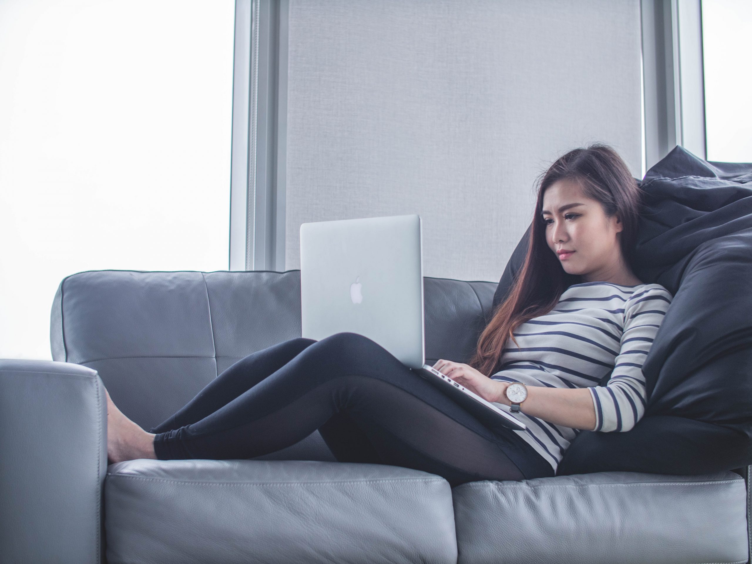 A young woman sits on a sofa wearing black trousers and a black and white striped top. She is working on a laptop