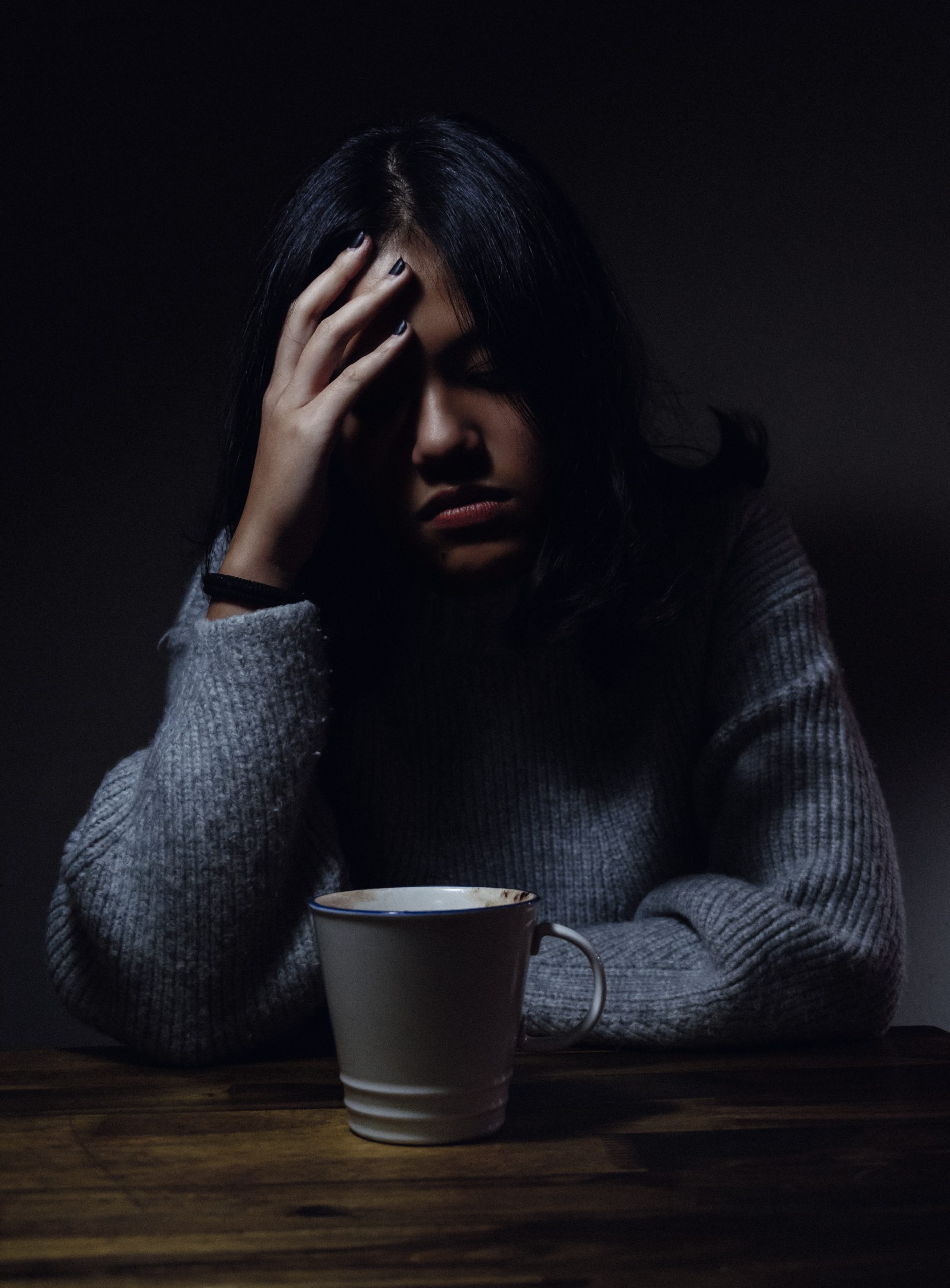 A young woman sits in a dark room with her head in her hands. She is leaning on a wooden table with a coffee mug in front of her.