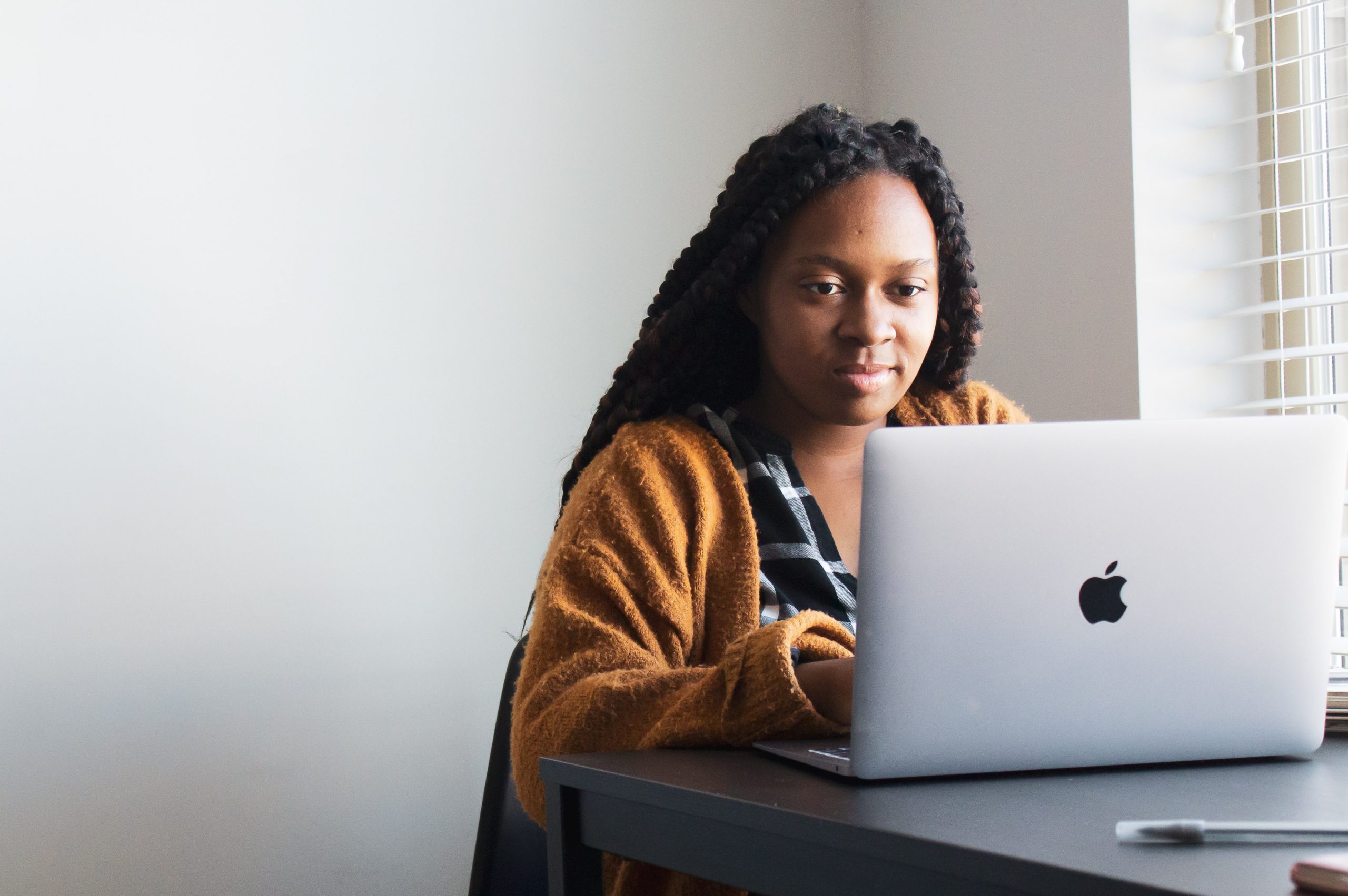A Black woman with dreads sits at a desk and writers on a MacBook. She is wearing a yellow cardigan.