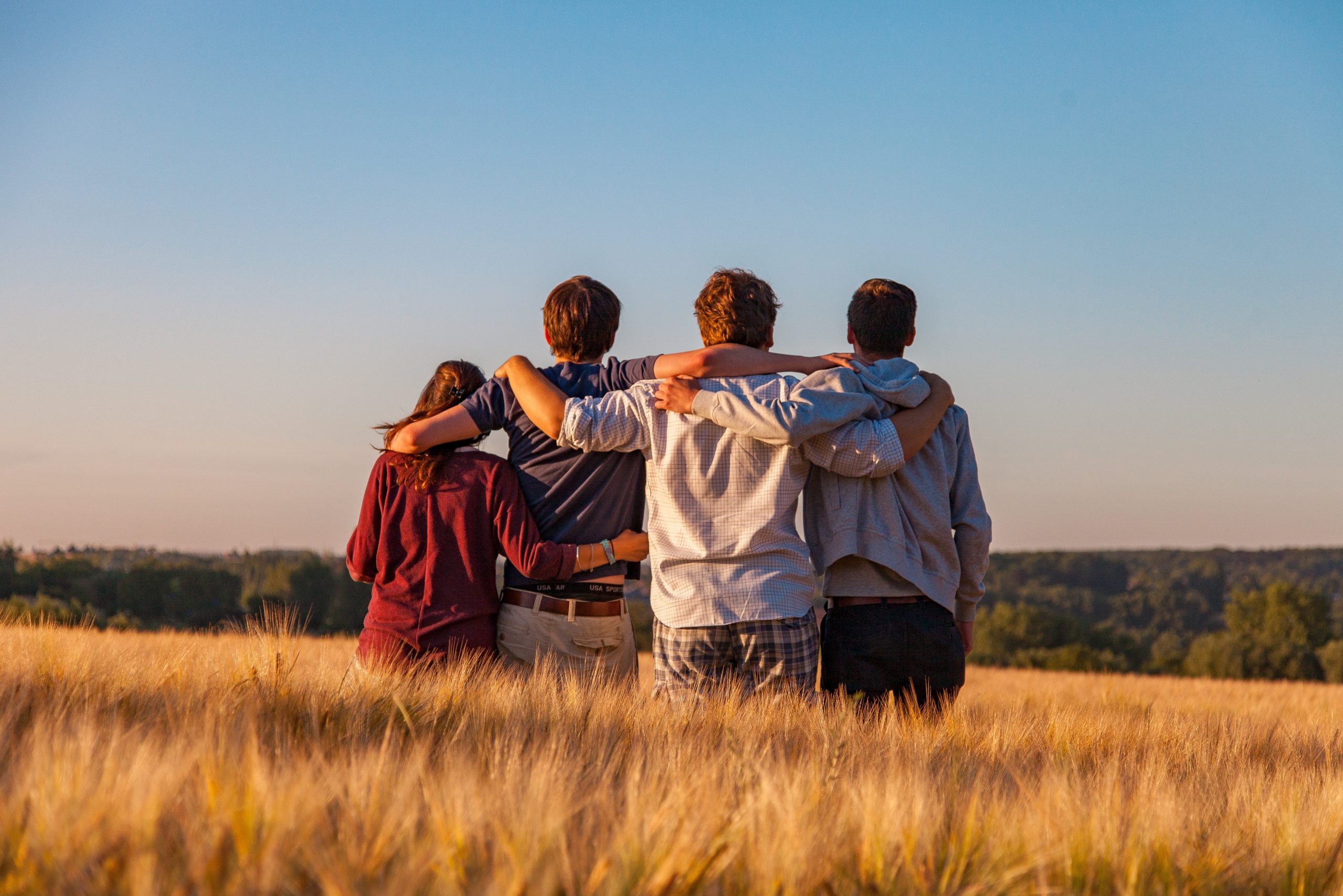 A group of young people stand looking out at a blue sky with their backs to the camera. They are all holding each other together.