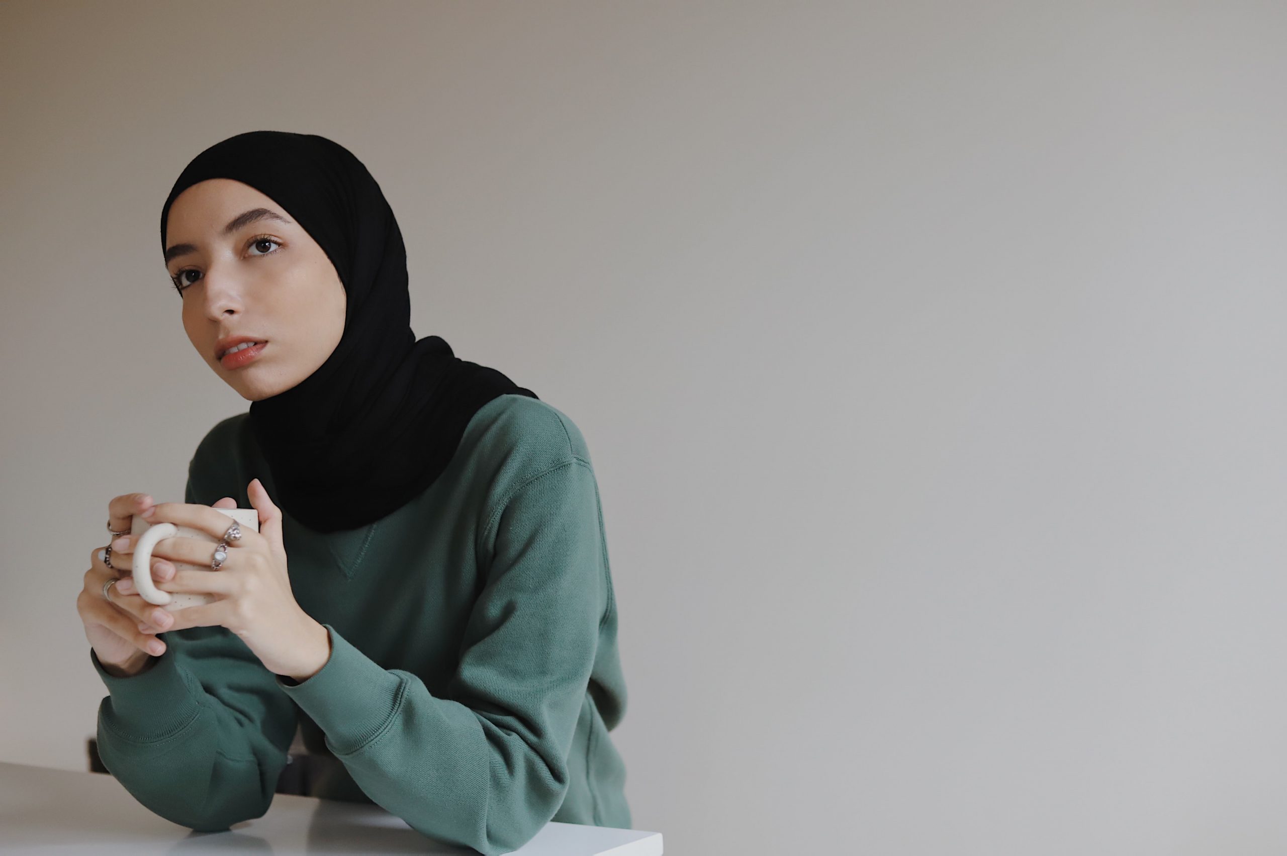 A young Muslim woman wearing a black hijab and green/blue jumper olds a mug of coffee and looks penseive.