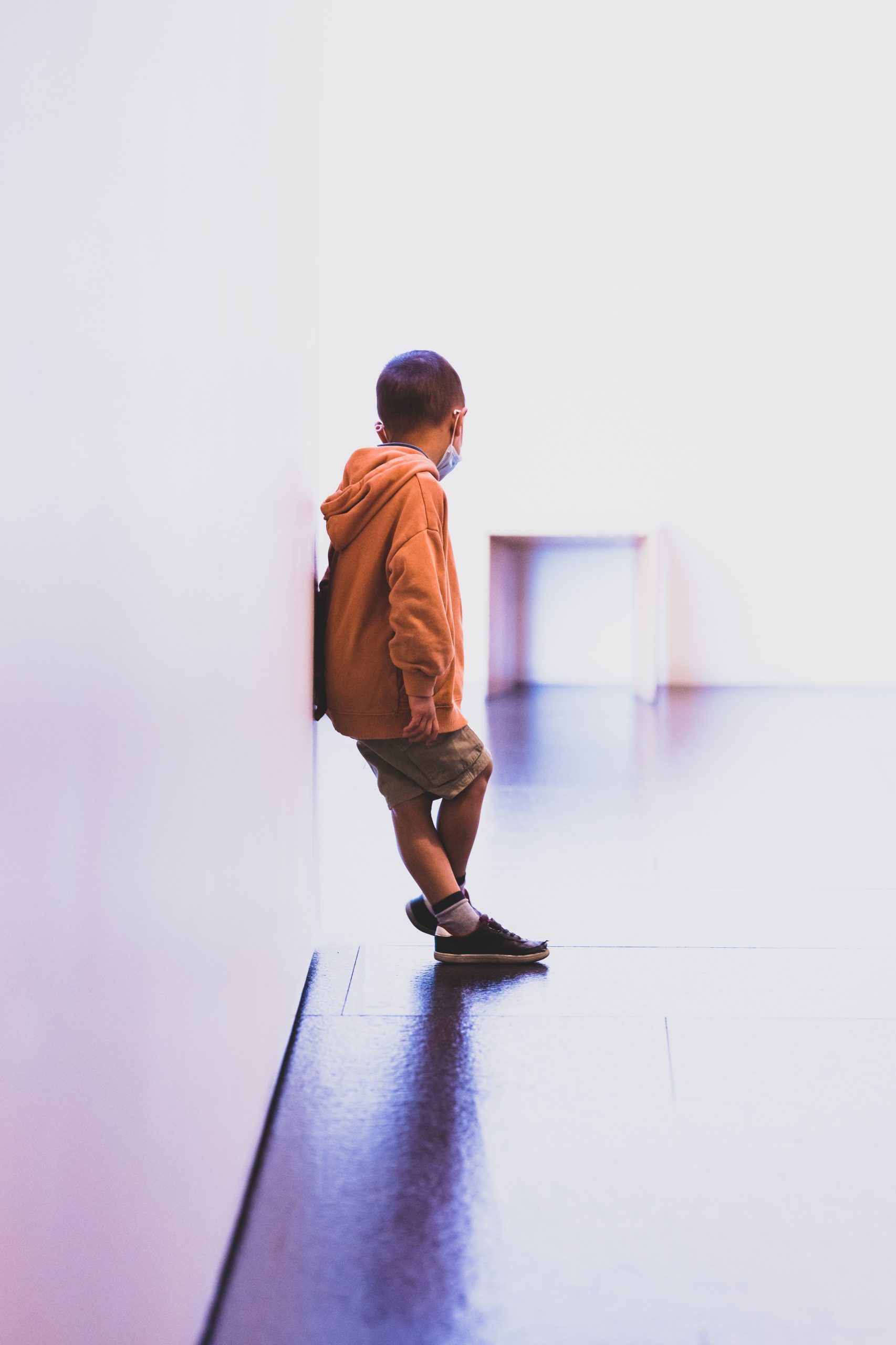 A young boy wearing an orange hoodie and khaki shorts leans agains a white wall and looks away from the camera