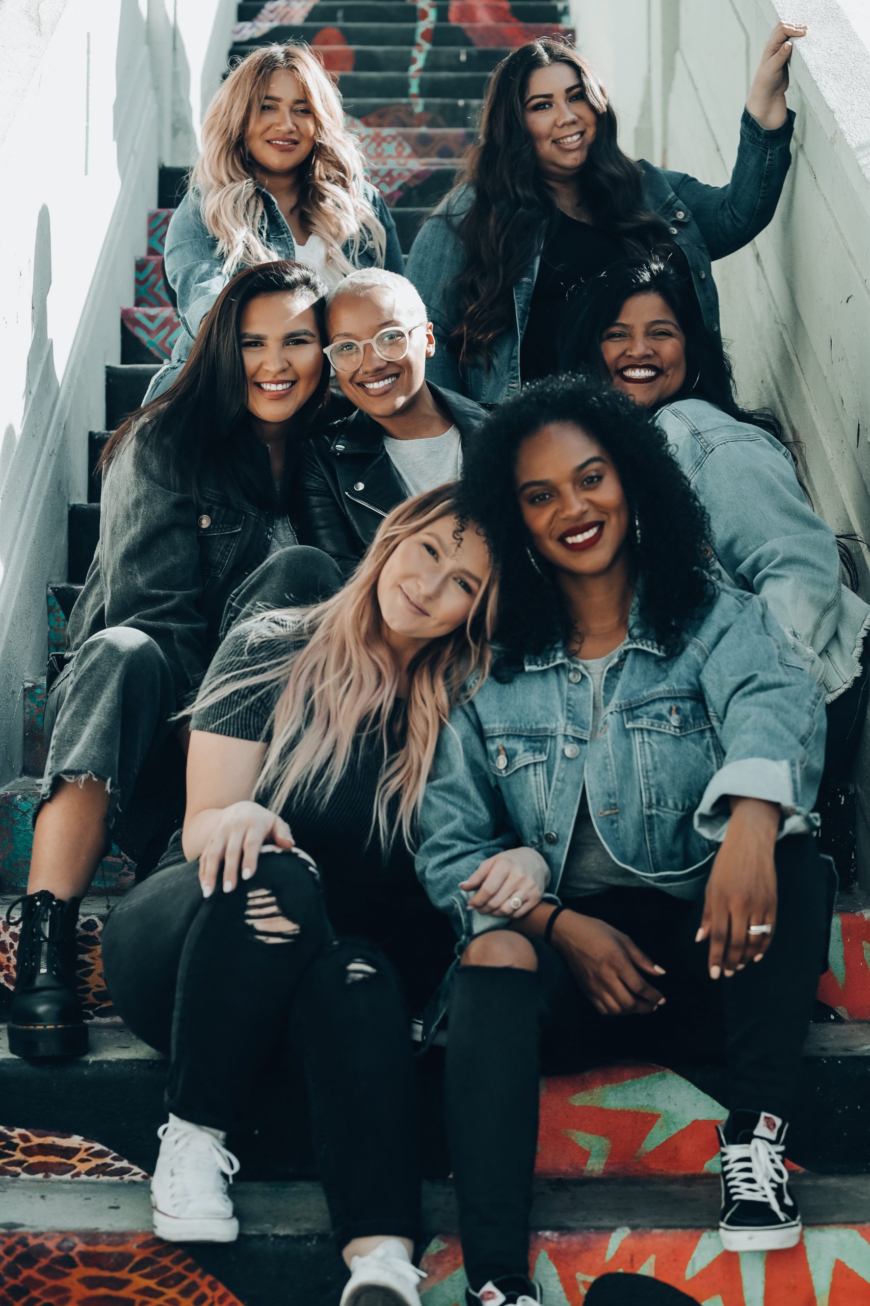 A grop of young women of all sizes and races sit on a flight of stairs smiling at the camera. They are all dressed casually and are smiling at the camera.