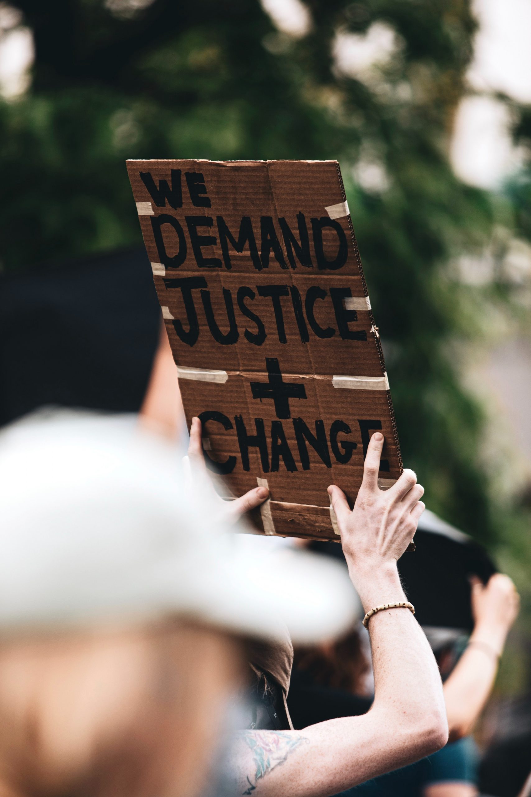 A person holds a sign aloft during a protest. Only their arms can be seen and the sign reads "we demand justice + change" in capital letters