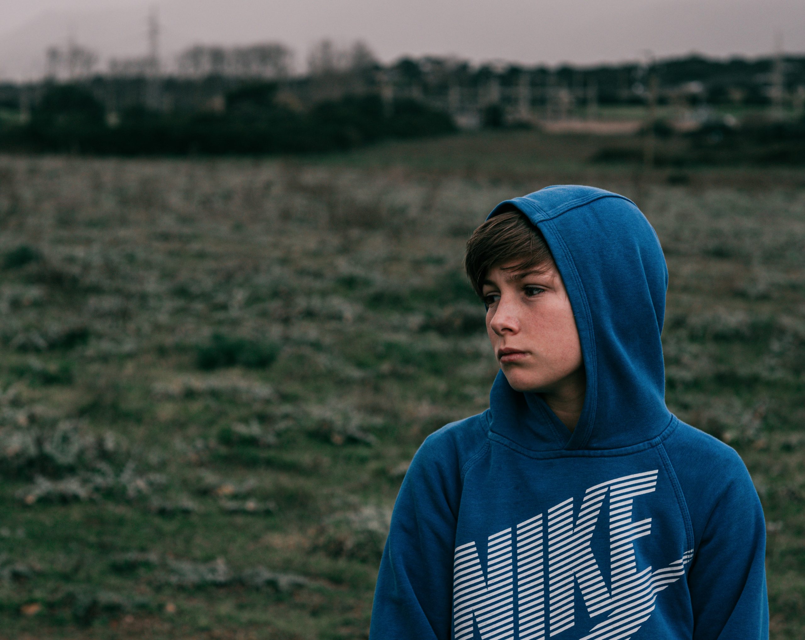 A young boy wearing a Nike hoodie in blue looks sad. He stands against a background of a green field at lowlight