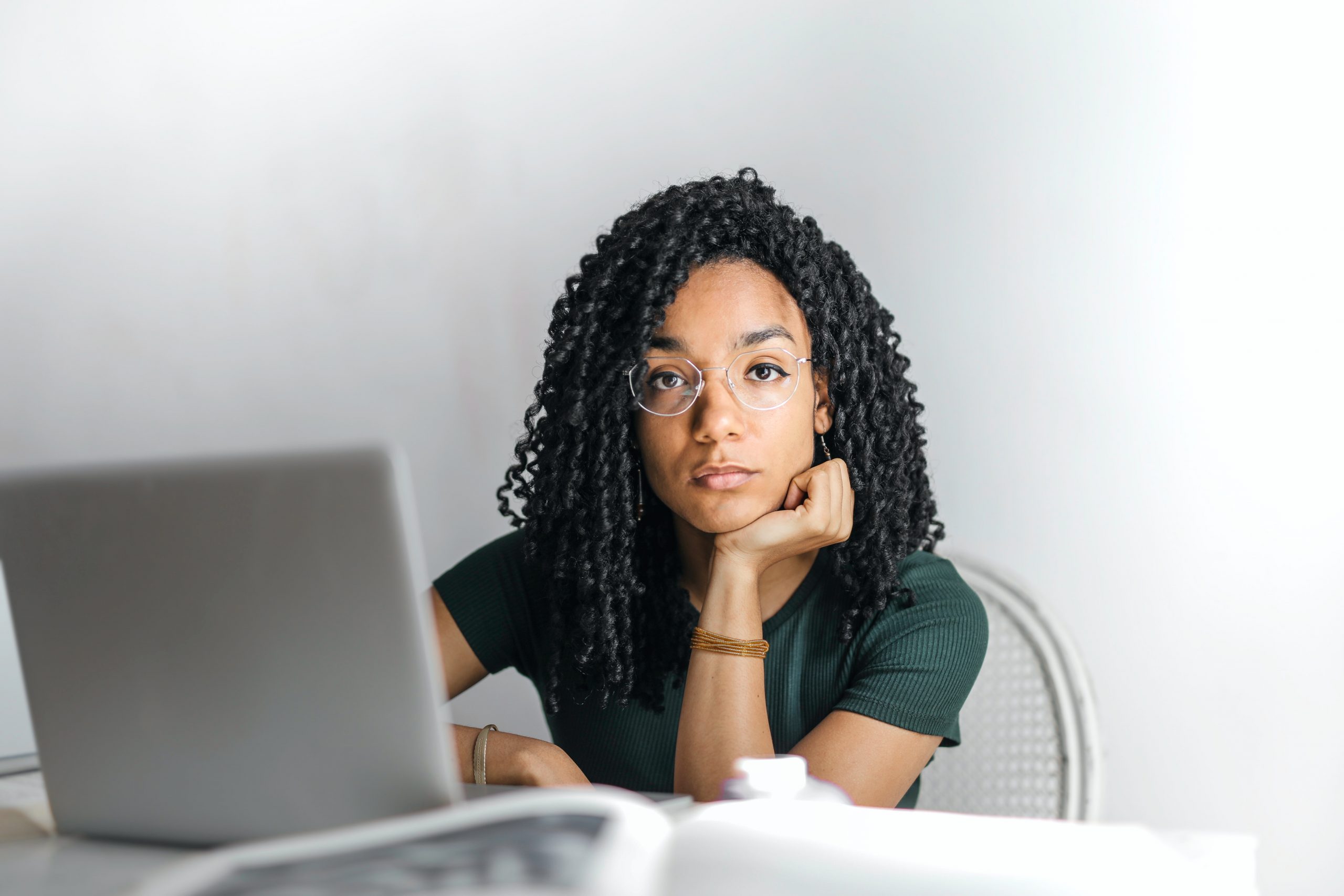 A young Black woman rests her chin on her palm and looks directly into the camera while a laptop sits open on the desk.