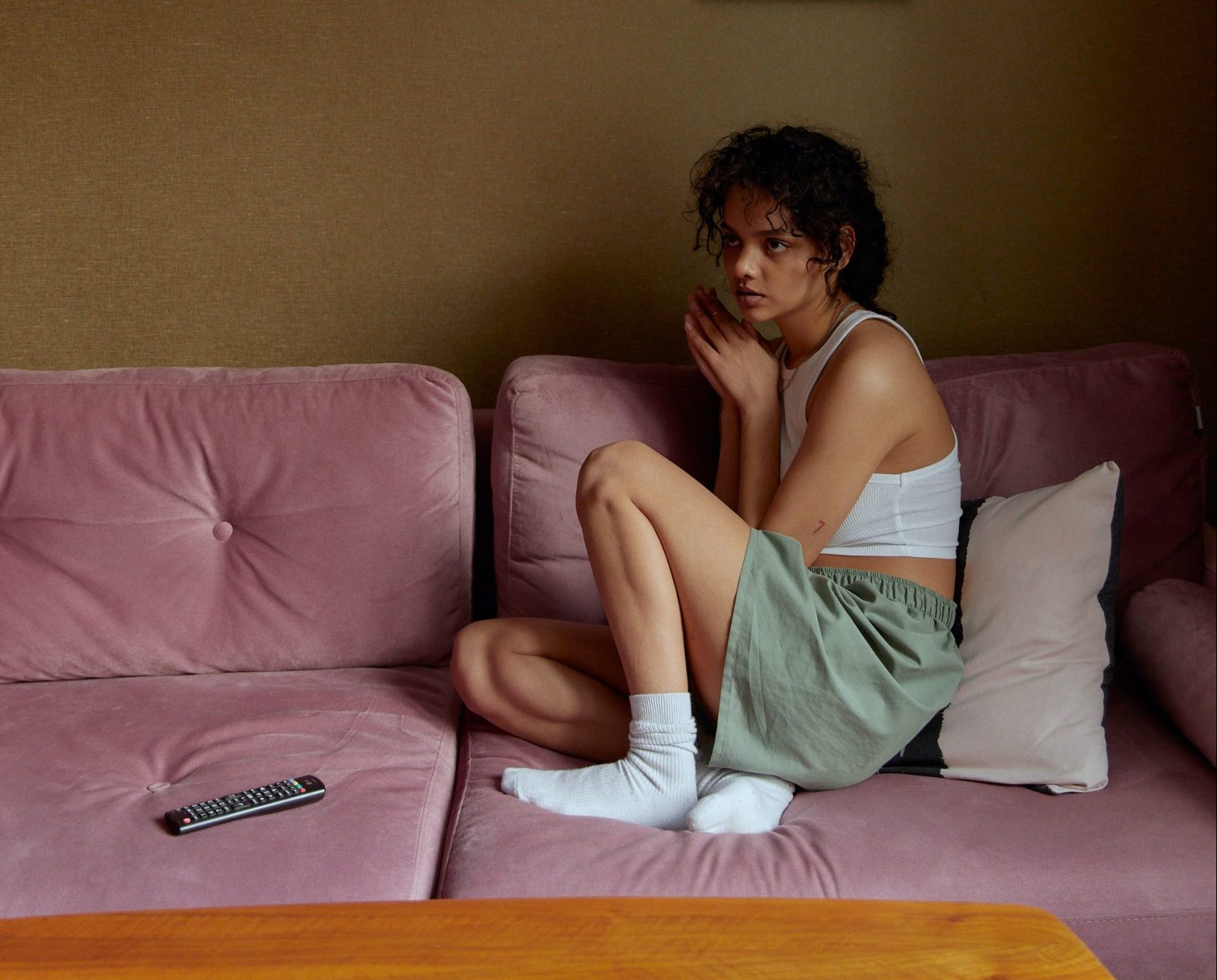 A young woman wears a crop top, shorts and white socks. She sits huddled up on a pink sofa looking pensieve.