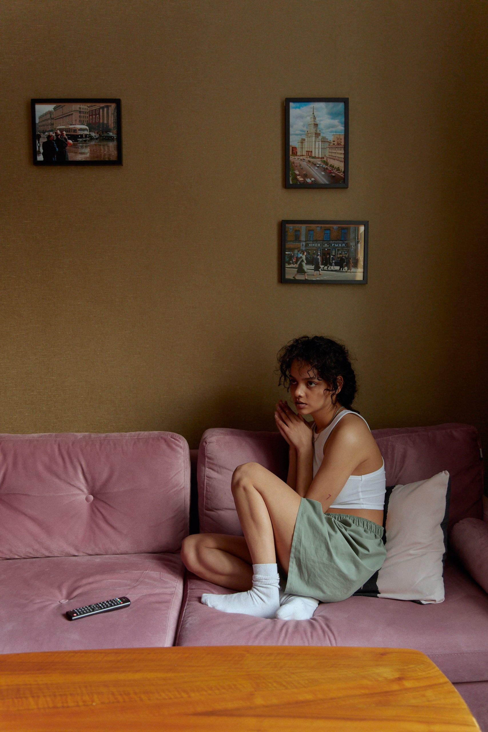 A young woman wears a crop top, shorts and white socks. She sits huddled up on a pink sofa looking pensieve.