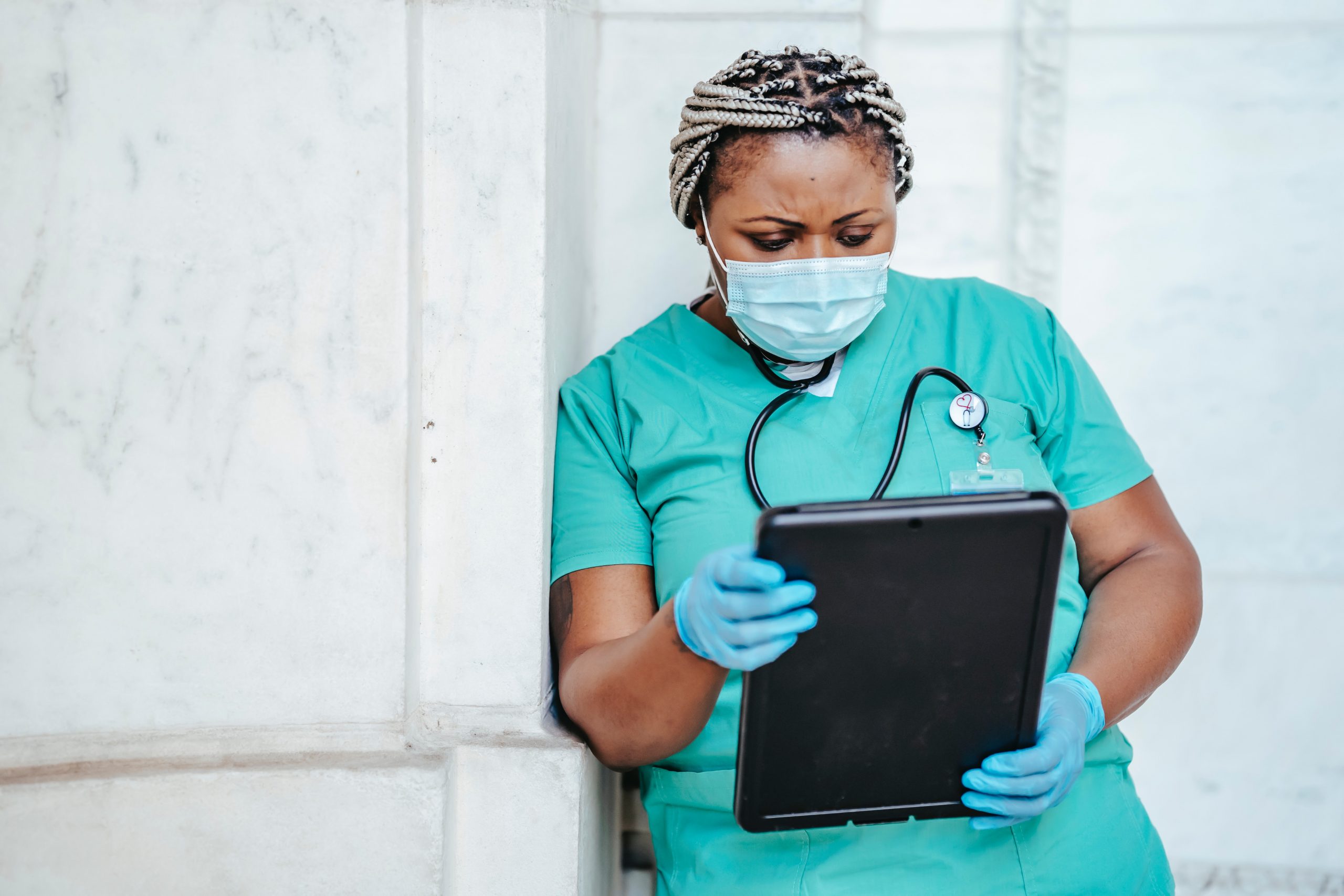 A Blac female doctor leans against a wall while reading a tablet. She wears a stethoscope, disposable gloves and a face mask. 