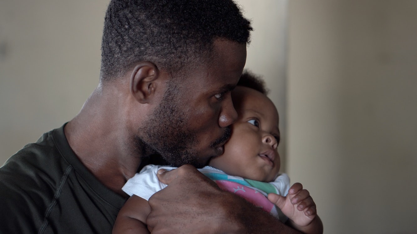 A young Black man holds his baby close to his face and kisses their cheek