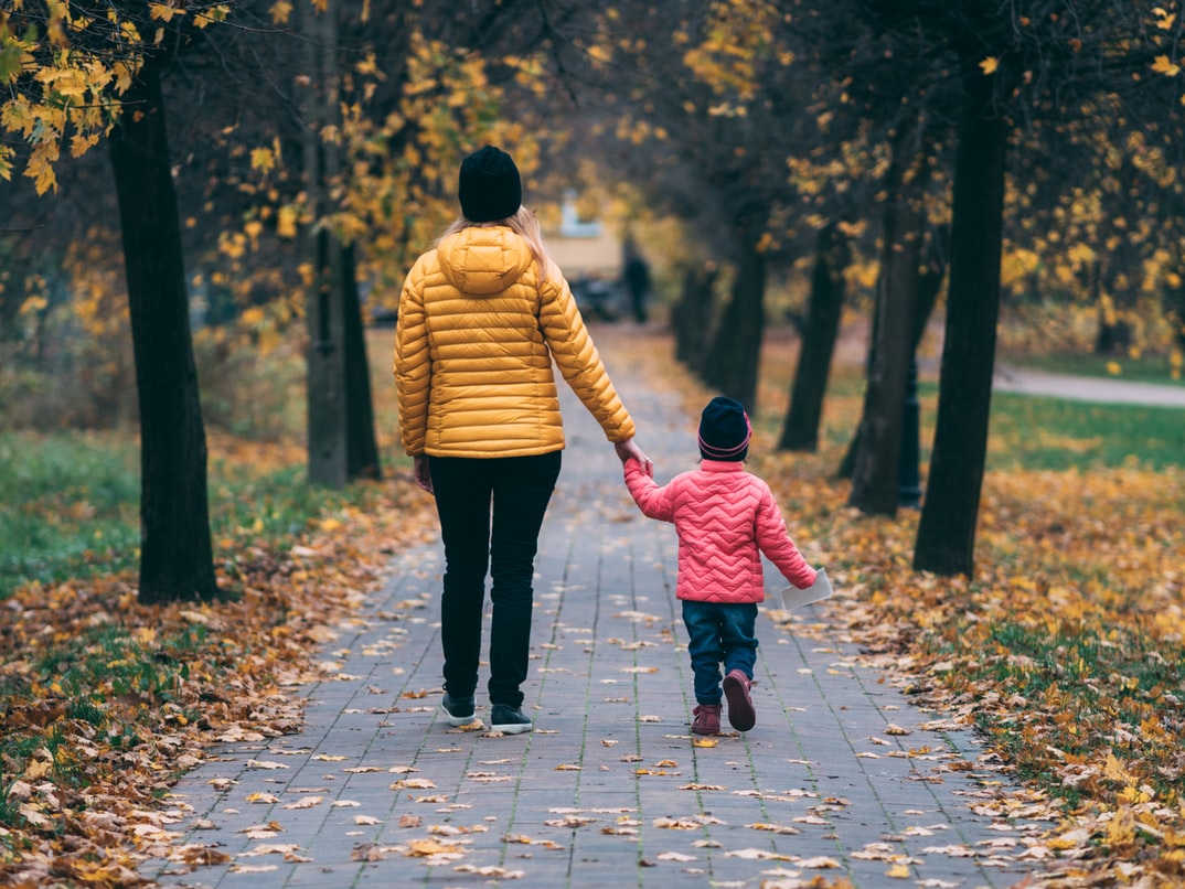 A young mother wearing a yellow jacke holds the hand of a small child wearing a pink jacket and walks away from the camera down a path between trees.