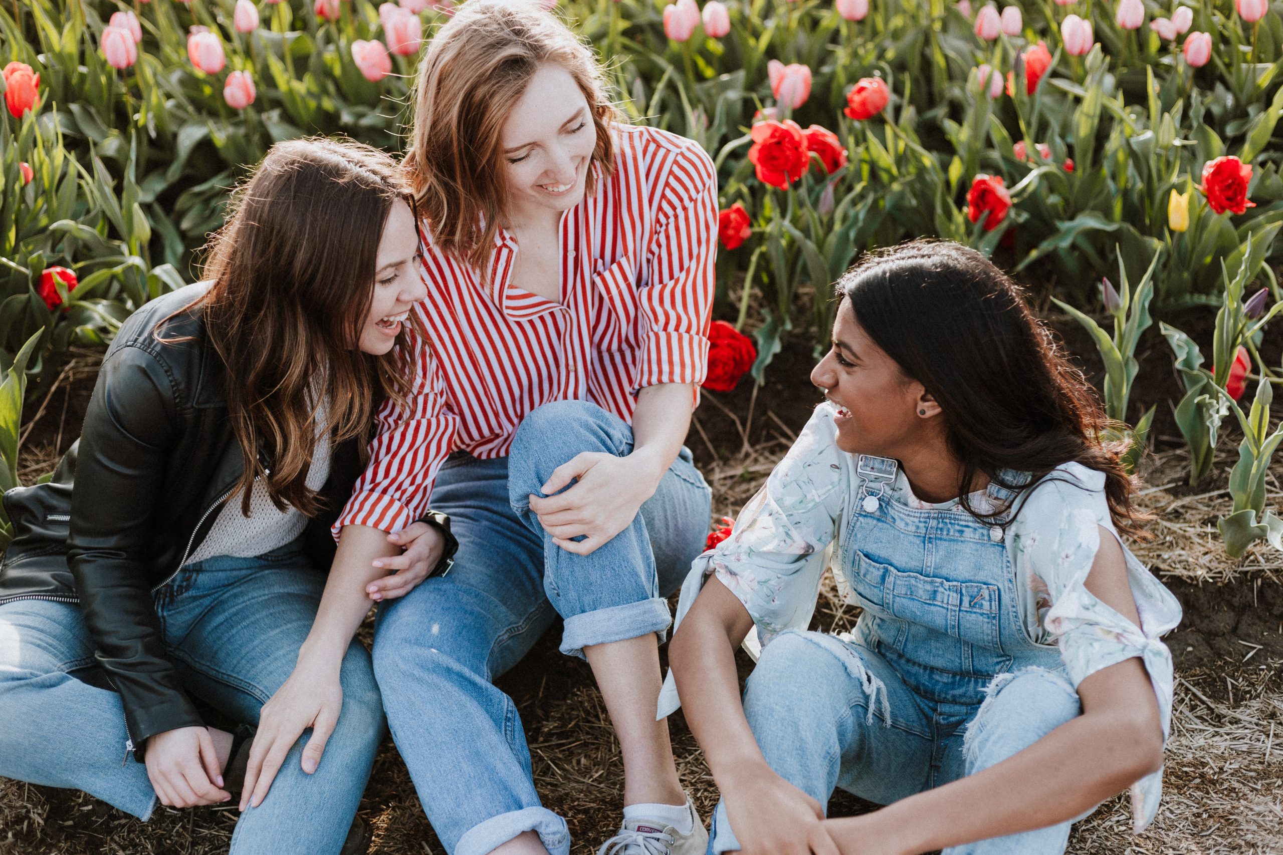 A group of three young women wearing jeans sit on the ground chatting and laughing