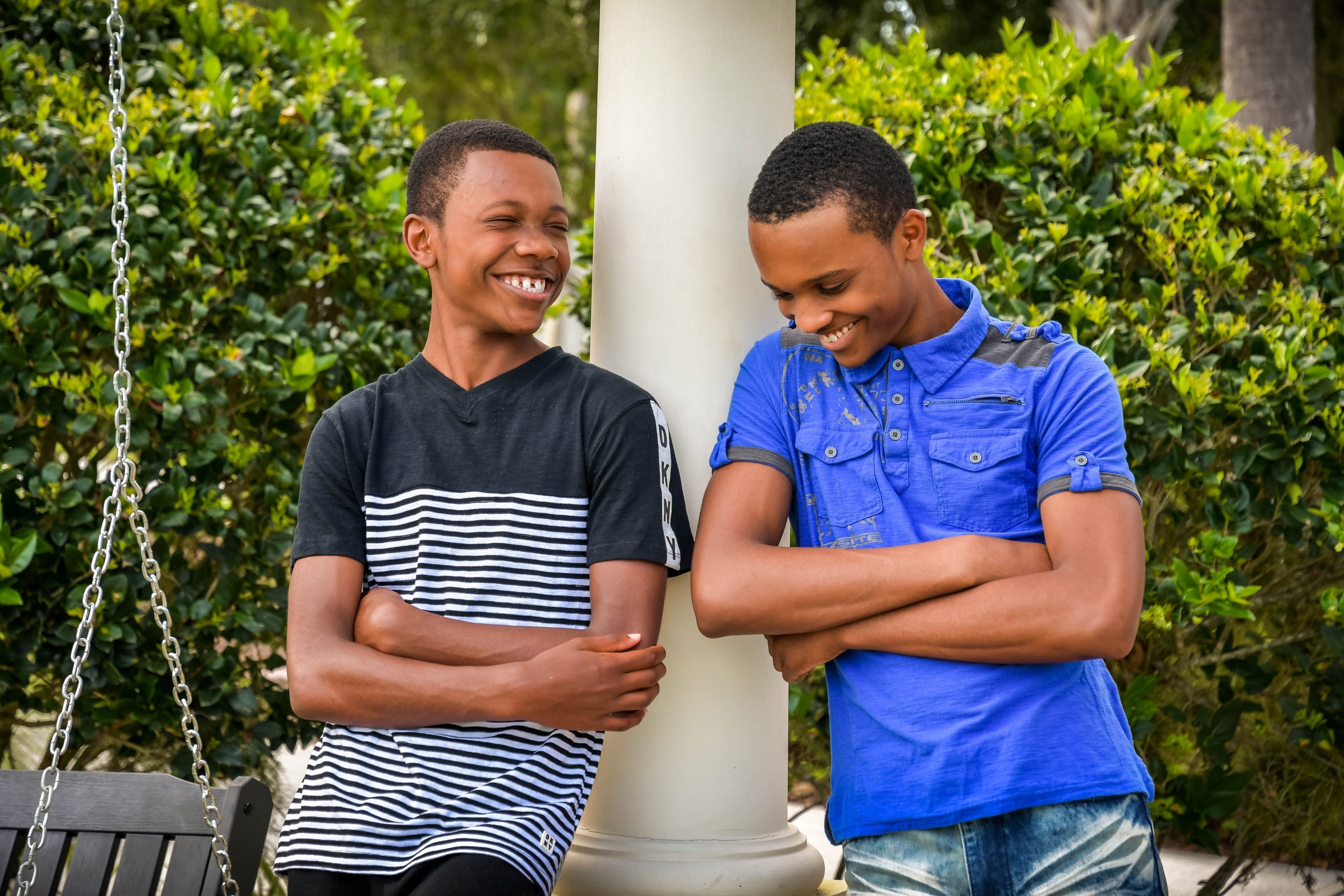 Two young black boys lean against a pole and smile and chat. The one on he left wears a navy and white t-shirt and the one on the right wears a bright blue one.