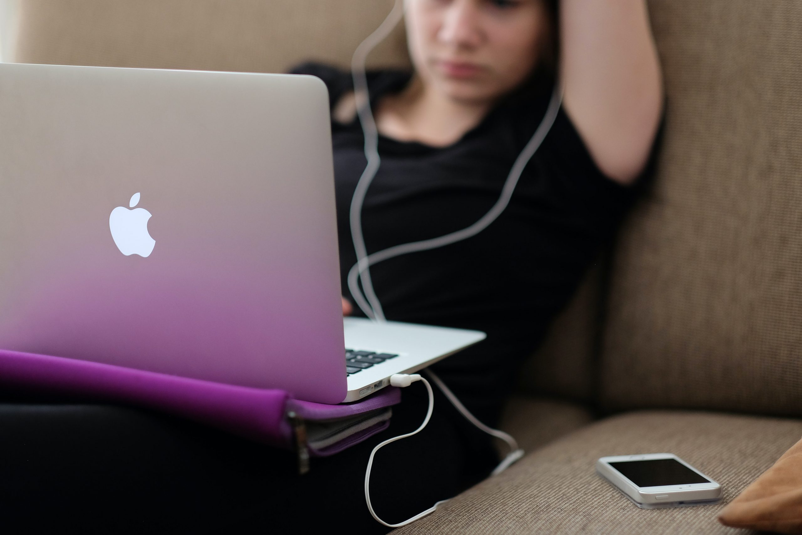 A young person sits on their laptop on a sofa. The shot shows them with a MacBook on their lap and a phone by their side. They are wearing earphones.