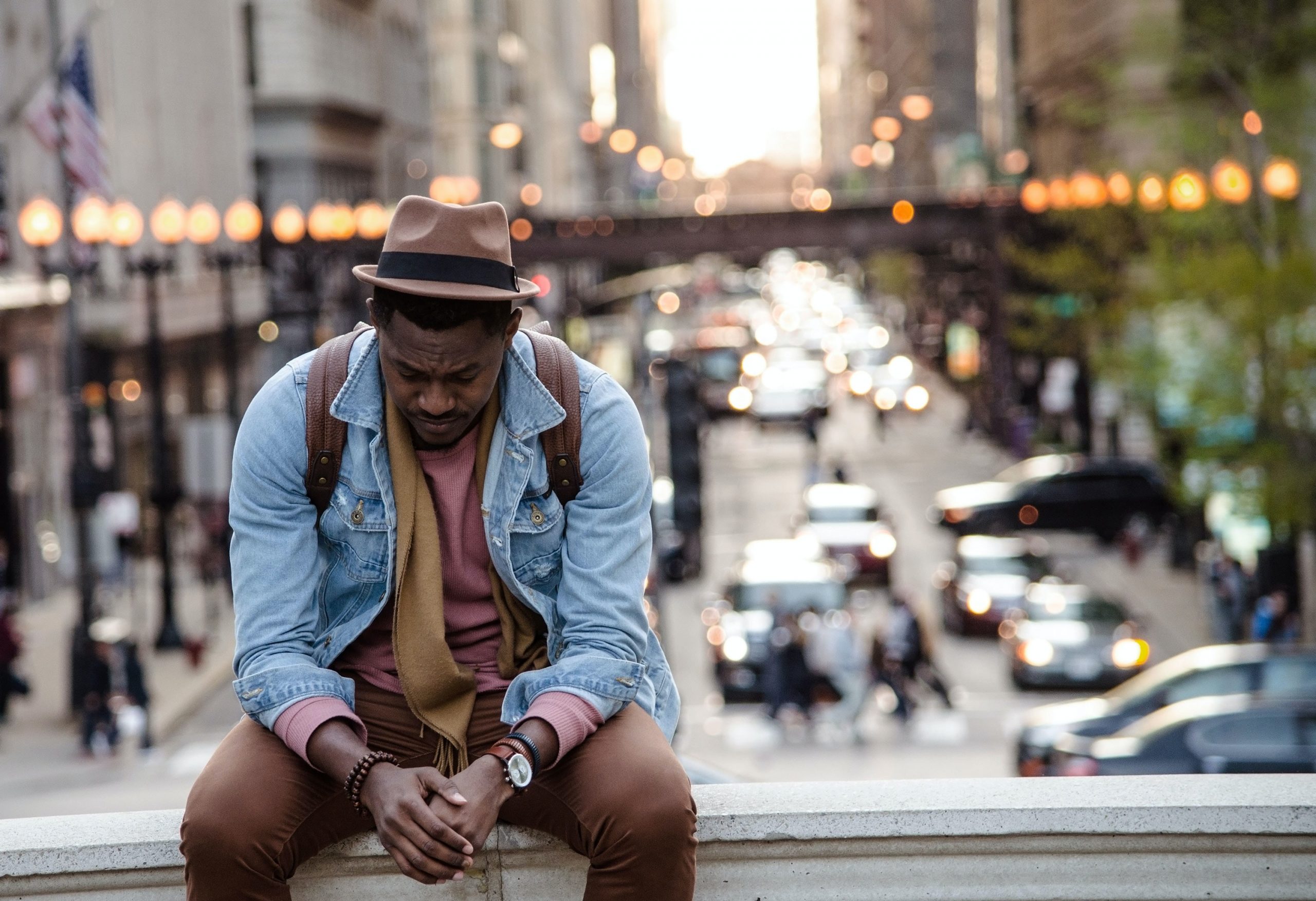 A young Black man sits on a ledge with his hands clasped looking contemplative. He wears a light blue denim jacket and maroon trousers.