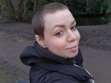 Alex Birch is a white woman and has shaved brown hair. She wears a black hoodie and looks back at the camera over her shoulder.
