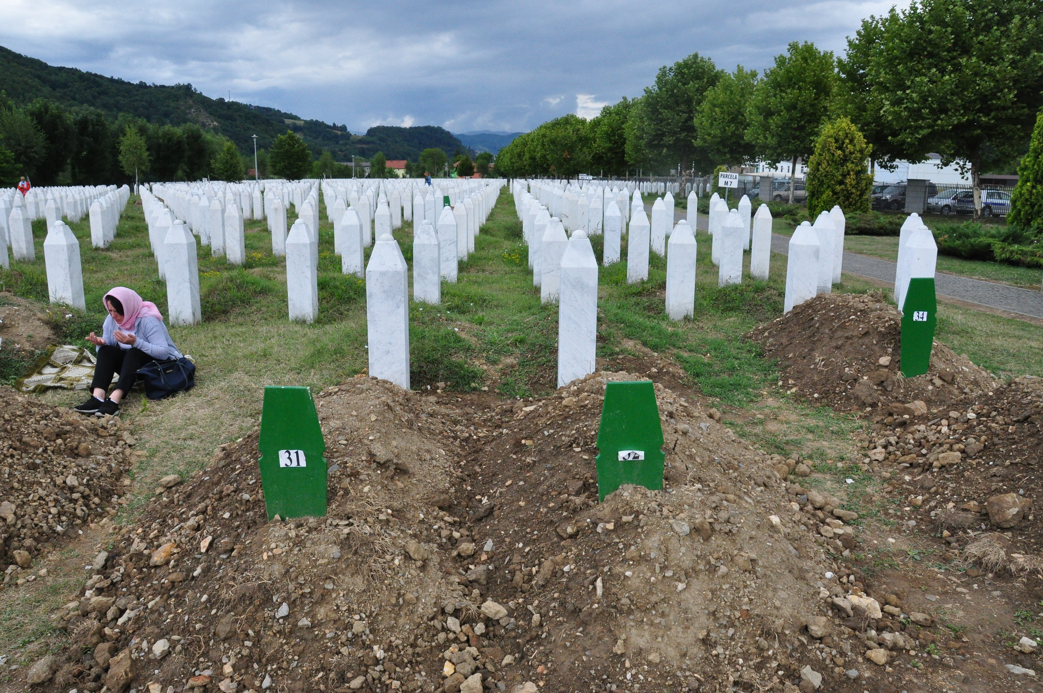 A grave site in Bosnia for victims of the genocide. In the foreground there are freshly dug graves with green markers on them and further back there is a field full of white stone grave markers